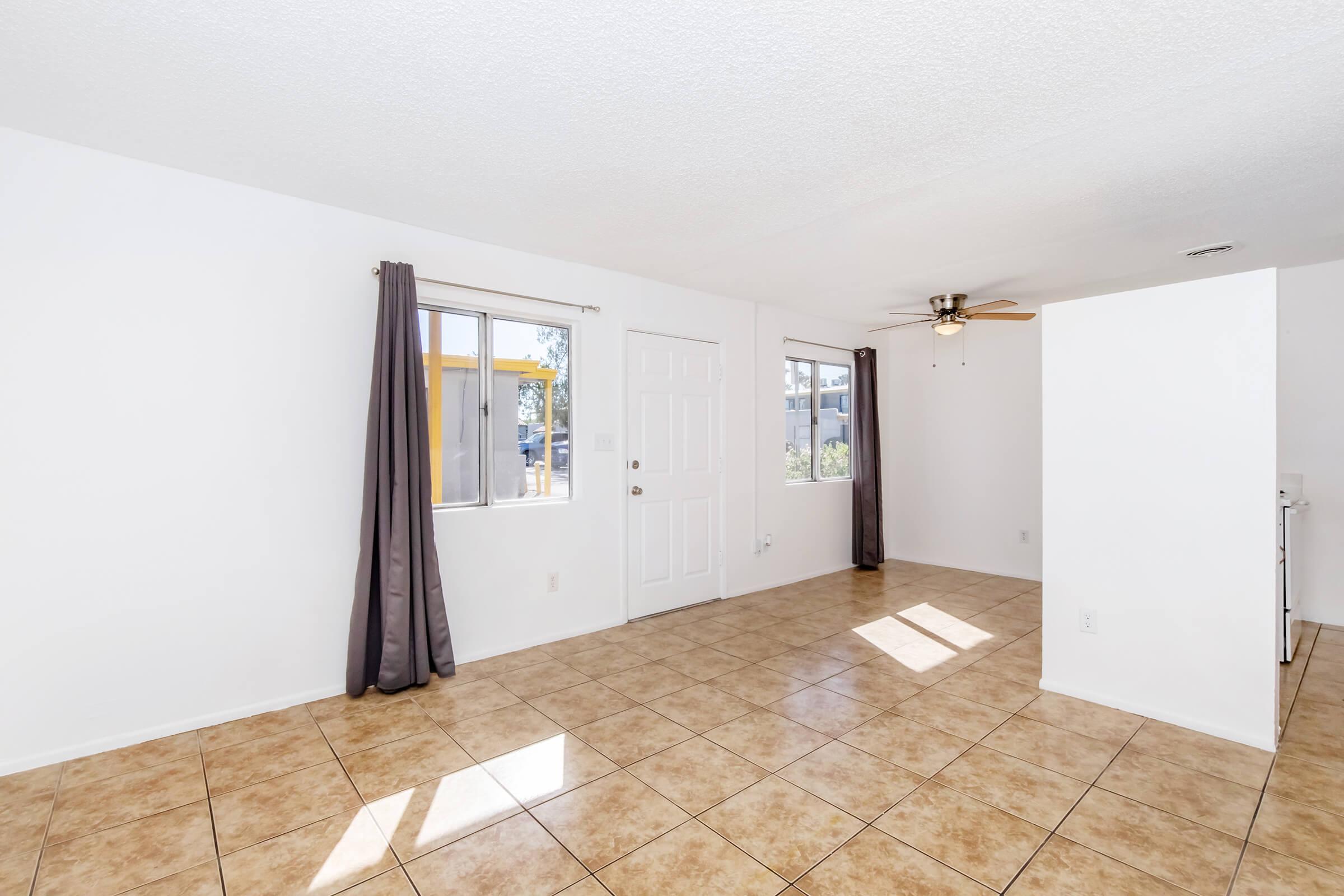 An interior view of a bright, clean living room featuring beige tiled floors, white walls, and a ceiling fan. There are large windows with gray curtains allowing natural light. A door leads outside, and the layout appears open with no visible furniture.