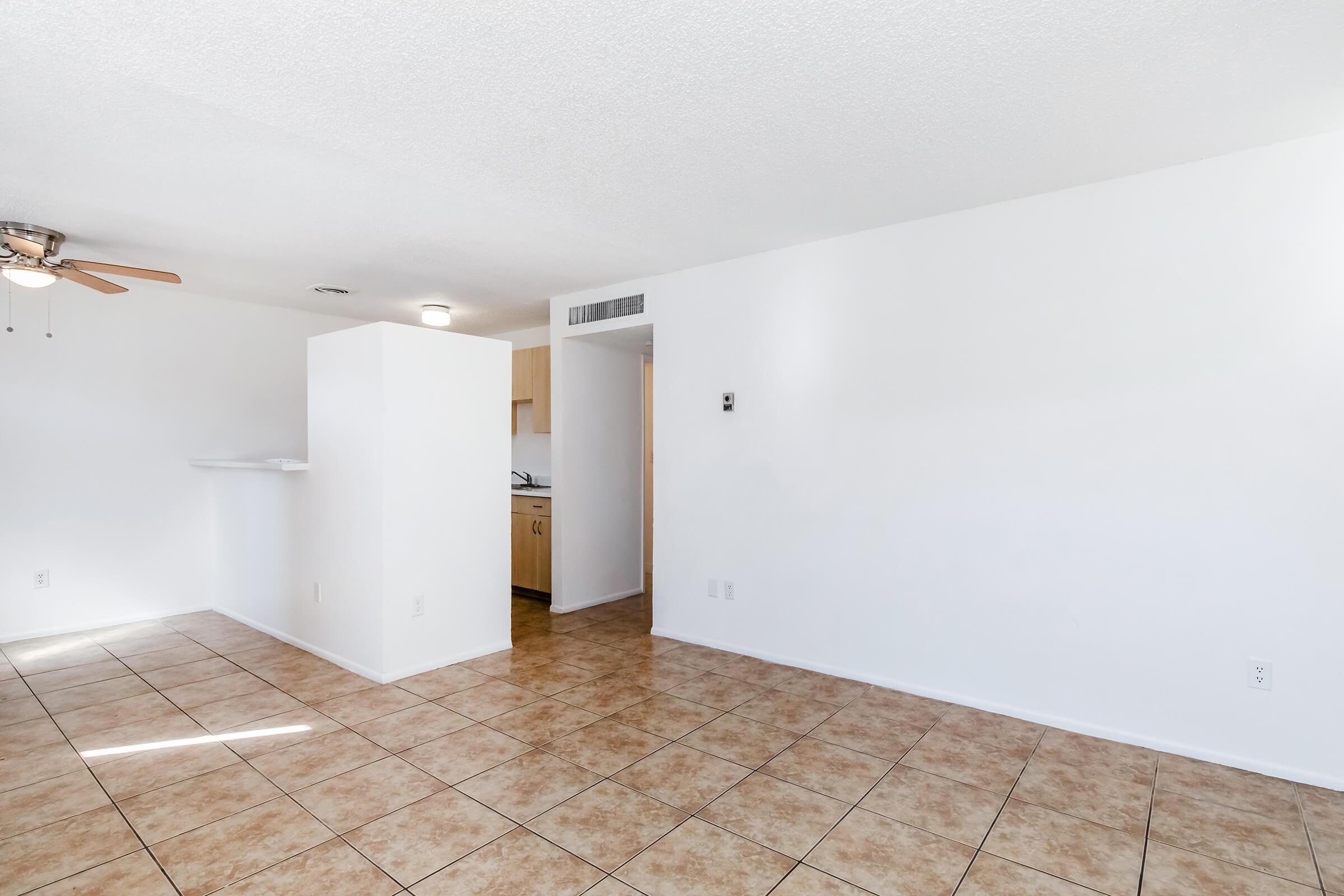 Empty room featuring light-colored walls, tiled flooring, and a ceiling fan. A small kitchen area is visible in the background, with an open layout leading to a doorway. The space is bright and airy, with natural light illuminating the room.