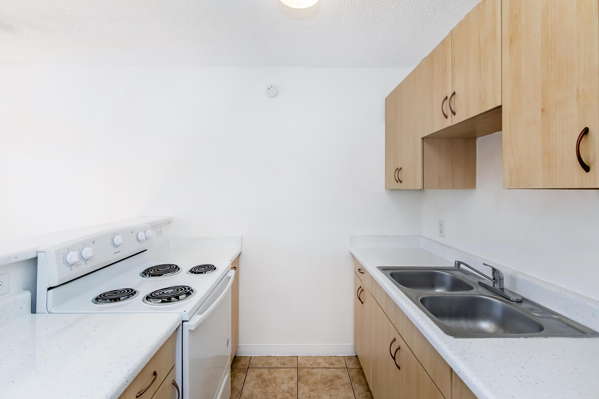 A bright, modern kitchen featuring white walls, wooden cabinetry, a stove with four burners, and a double sink. The countertops are light-colored with a speckled texture, and the floor has square tiles. The space is uncluttered and well-lit, creating a clean and inviting atmosphere.