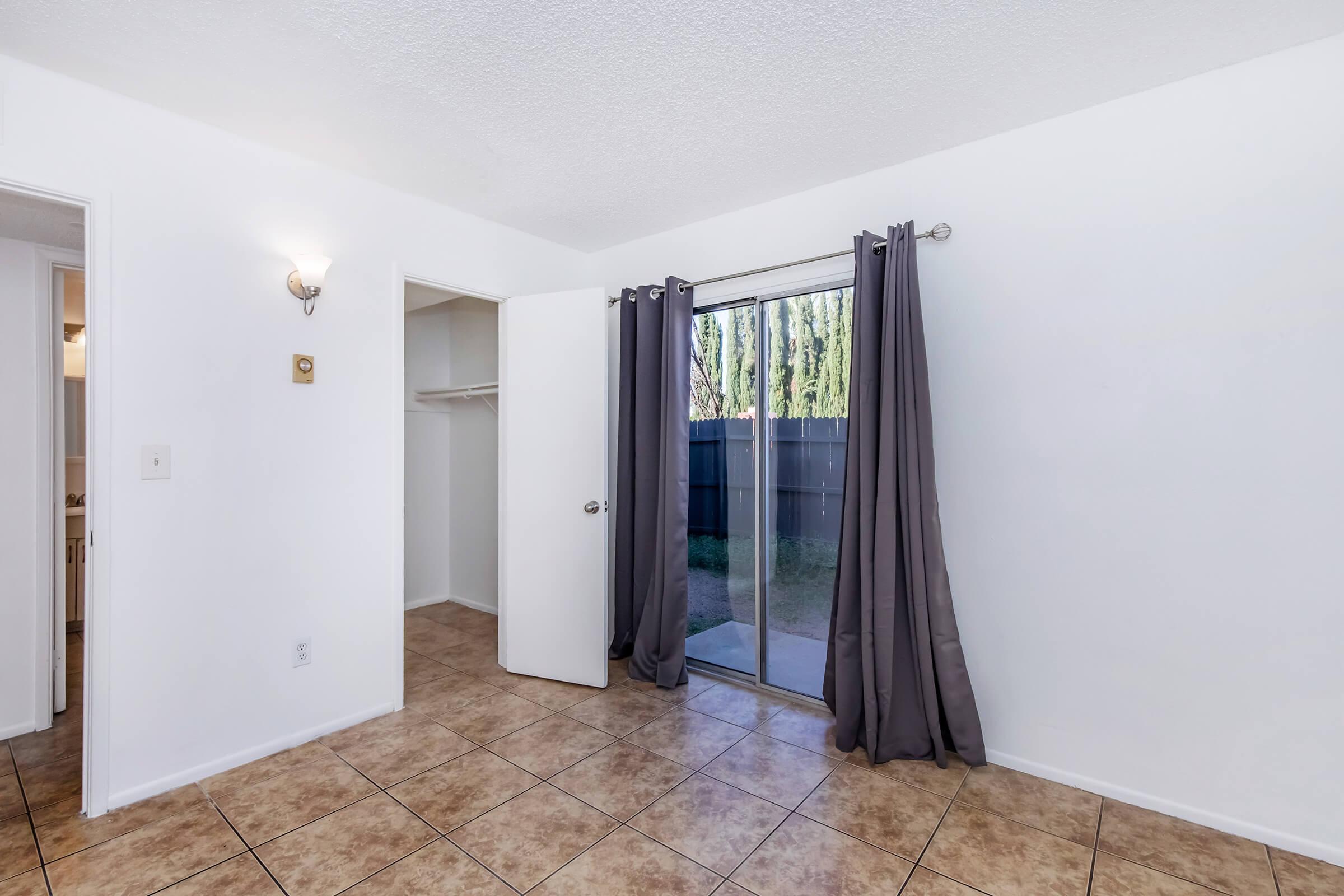 A small, well-lit room with white walls and tiled flooring. There is a sliding glass door with gray curtains leading to an outdoor area. To the left, a door leads to a closet. The space appears clean and minimalistic, suitable for various uses such as a bedroom or office.