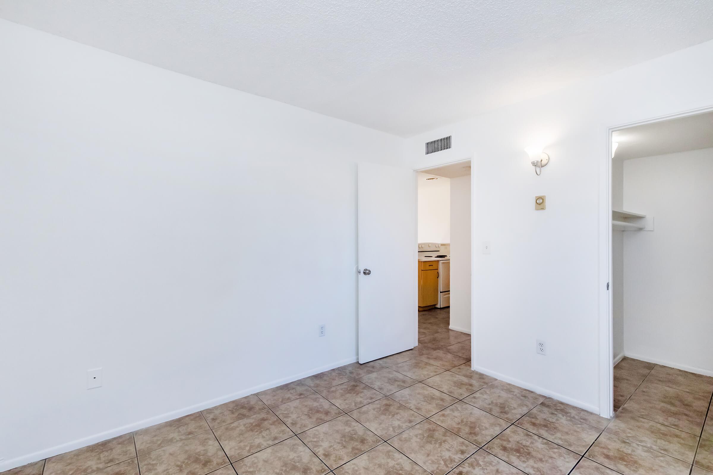 Empty room with white walls and tiled flooring. A door is visible on the left leading to another area. A small closet opening is seen on the right, and a light fixture is mounted on the wall. The room is well-lit, giving a clean and spacious appearance.