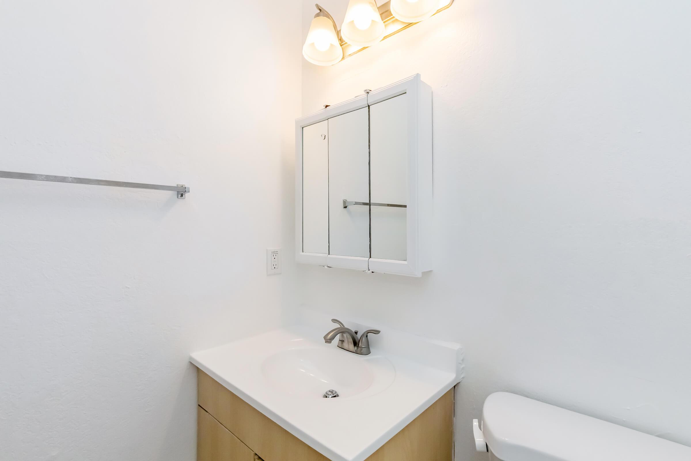 Modern bathroom interior featuring a white sink with a faucet, a light-colored cabinet beneath, a mirrored medicine cabinet on the wall, and a toilet to the right. Soft lighting from three light fixtures above adds a bright ambiance against the plain white walls.