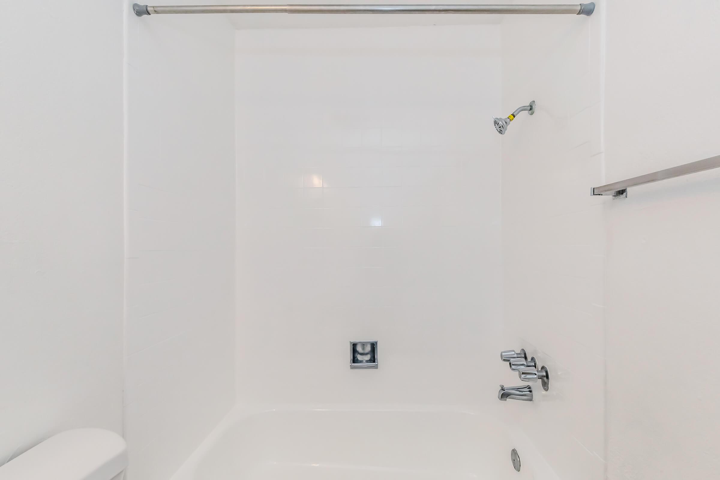 A clean, empty bathroom shower space featuring white tiled walls, a bathtub, and a showerhead. A wall-mounted towel bar is visible on one side, and a drain is located on the floor. The overall aesthetic is bright and minimalistic.