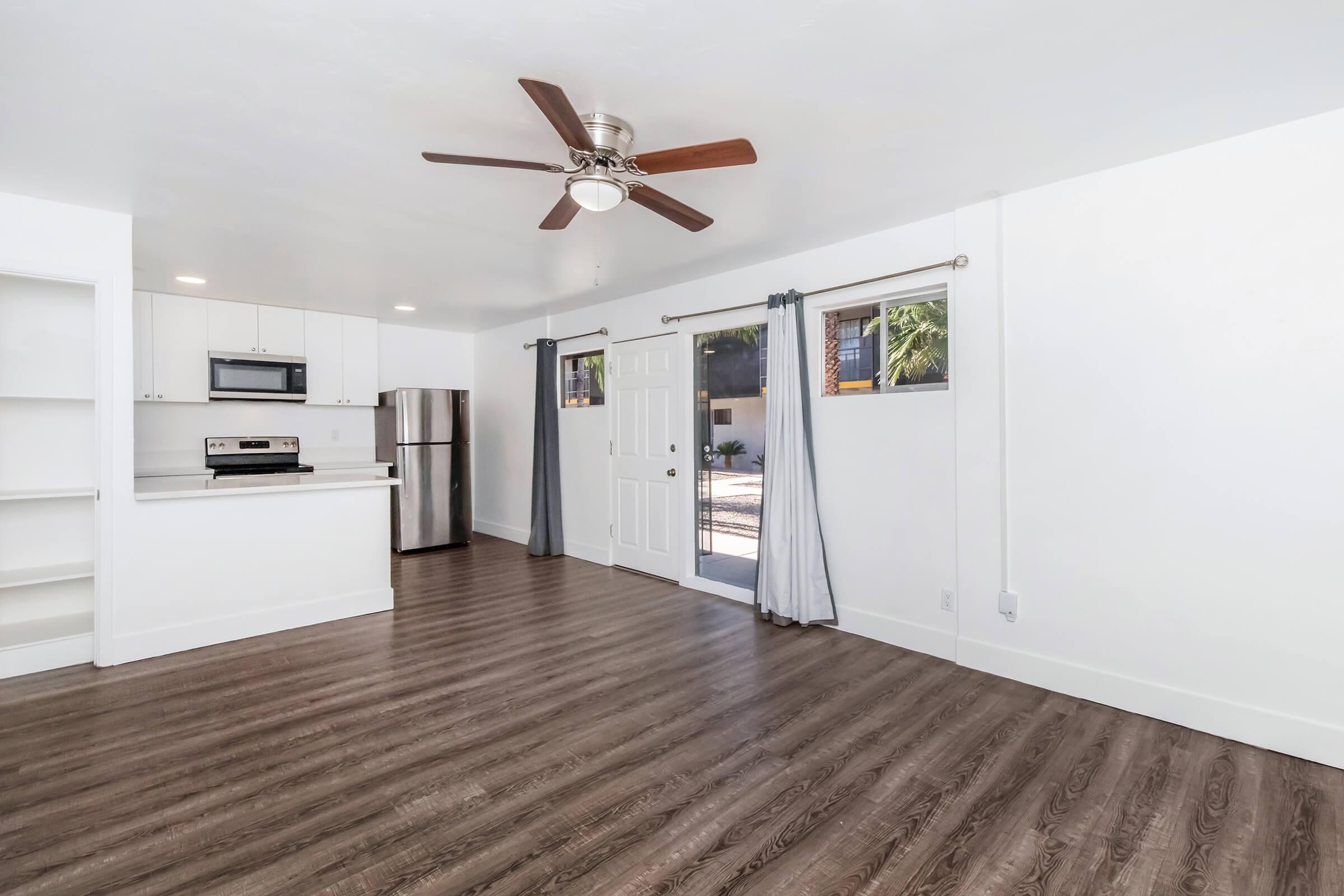 A bright, open living space with a modern kitchen featuring stainless steel appliances, a ceiling fan, and large windows. The walls are painted white, and the flooring is a rich, dark wood laminate. There are sliding glass doors leading outside, and light curtains frame the windows.