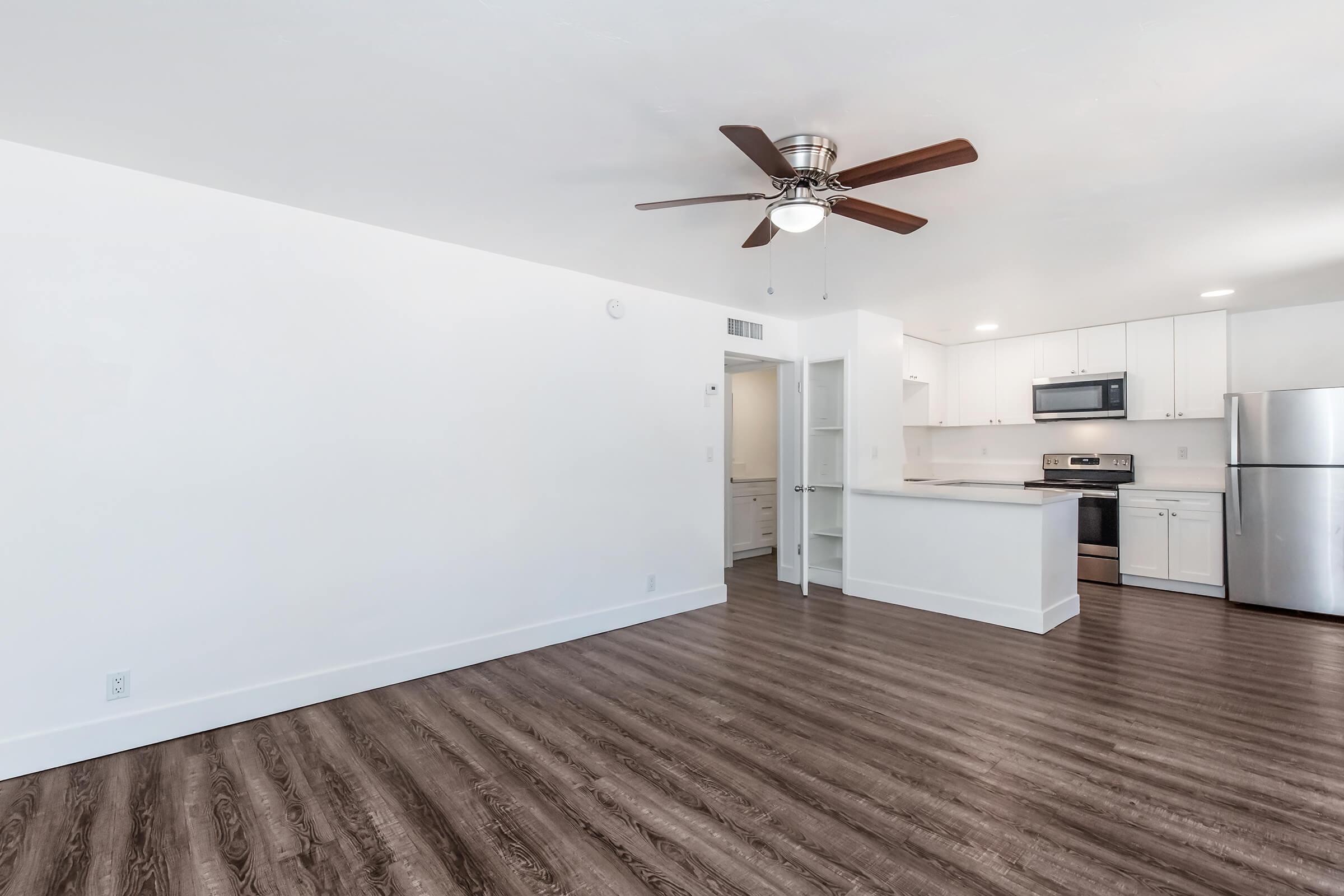 Interior of a modern, spacious living area featuring light-colored walls, a ceiling fan, and wood-like flooring. The kitchen is visible in the background with stainless steel appliances, including a refrigerator and oven, and white cabinets. The open layout creates a bright and inviting atmosphere.