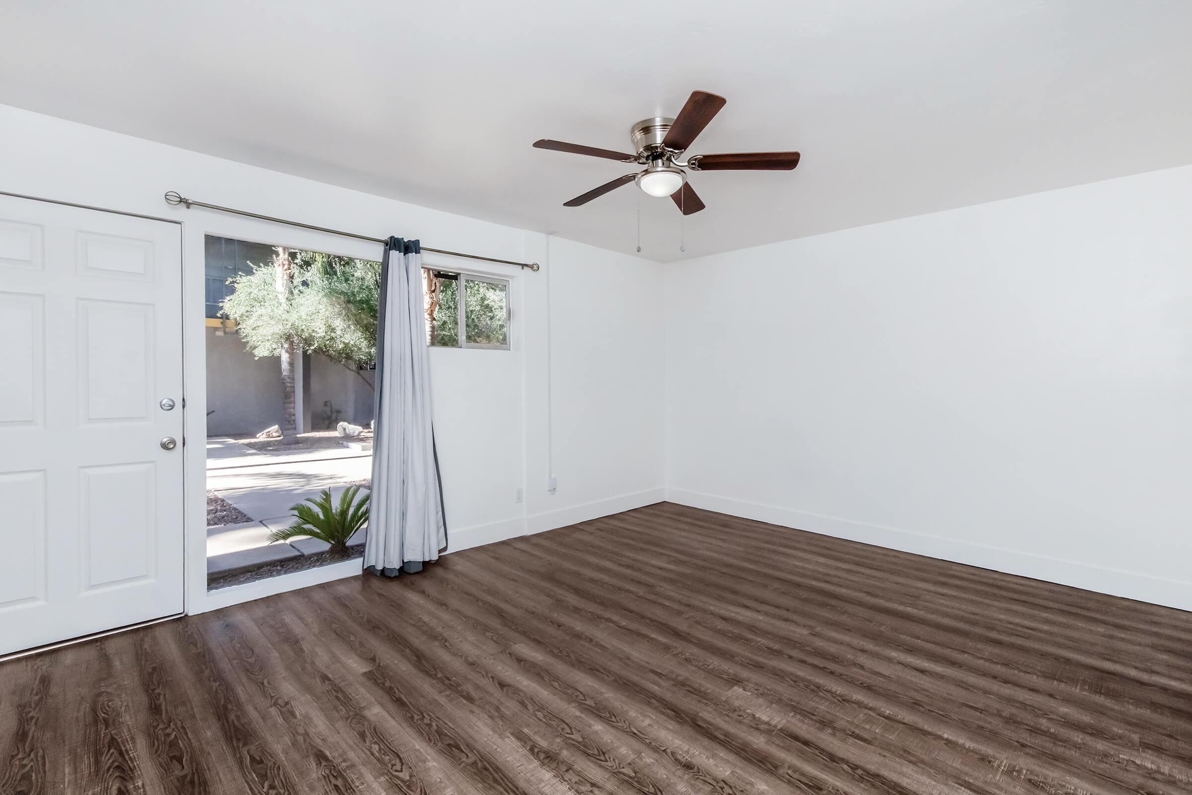 Bright and airy room featuring light wood flooring, a ceiling fan, and large windows with views of a garden. The walls are painted white, enhancing the spacious feel. A door leads outside, and curtains frame the window, adding a touch of softness to the modern aesthetic.