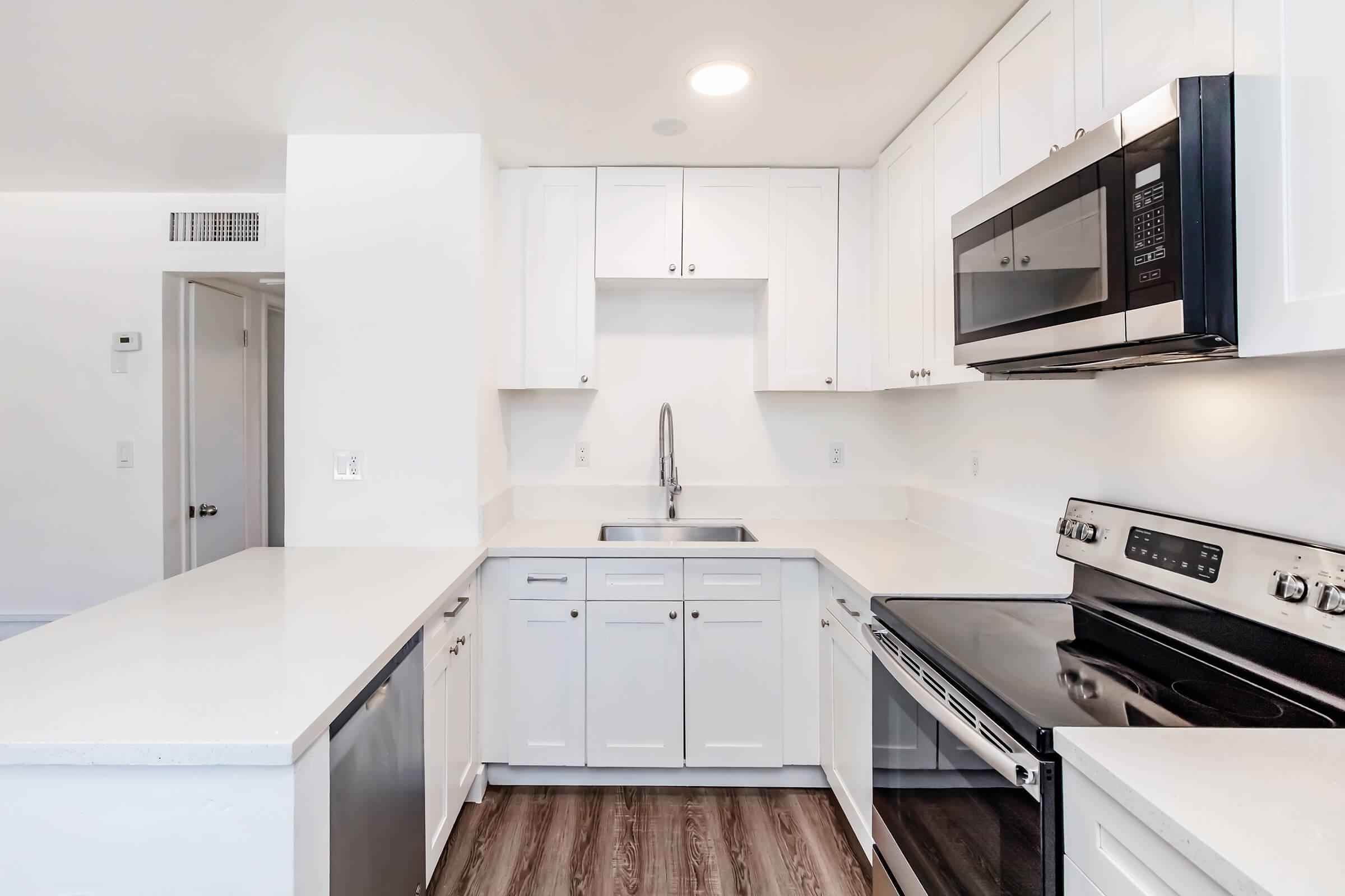 Modern kitchen featuring white cabinetry, a stainless steel microwave, an oven, and a dishwasher. The space has a sleek countertop and a single-basin sink, with light wood flooring and ample natural light. The layout is open and clean, creating a spacious atmosphere.