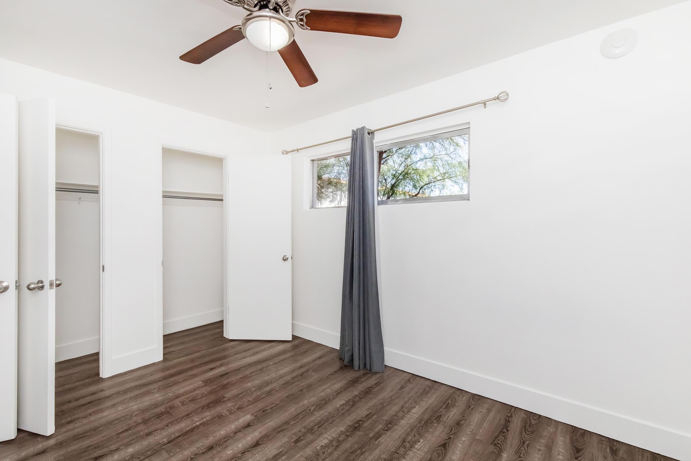 A bright, empty bedroom featuring a ceiling fan and large windows. Two open closet doors are visible, and a gray curtain hangs beside the window. The room has wooden flooring and white walls, creating a clean and modern look.