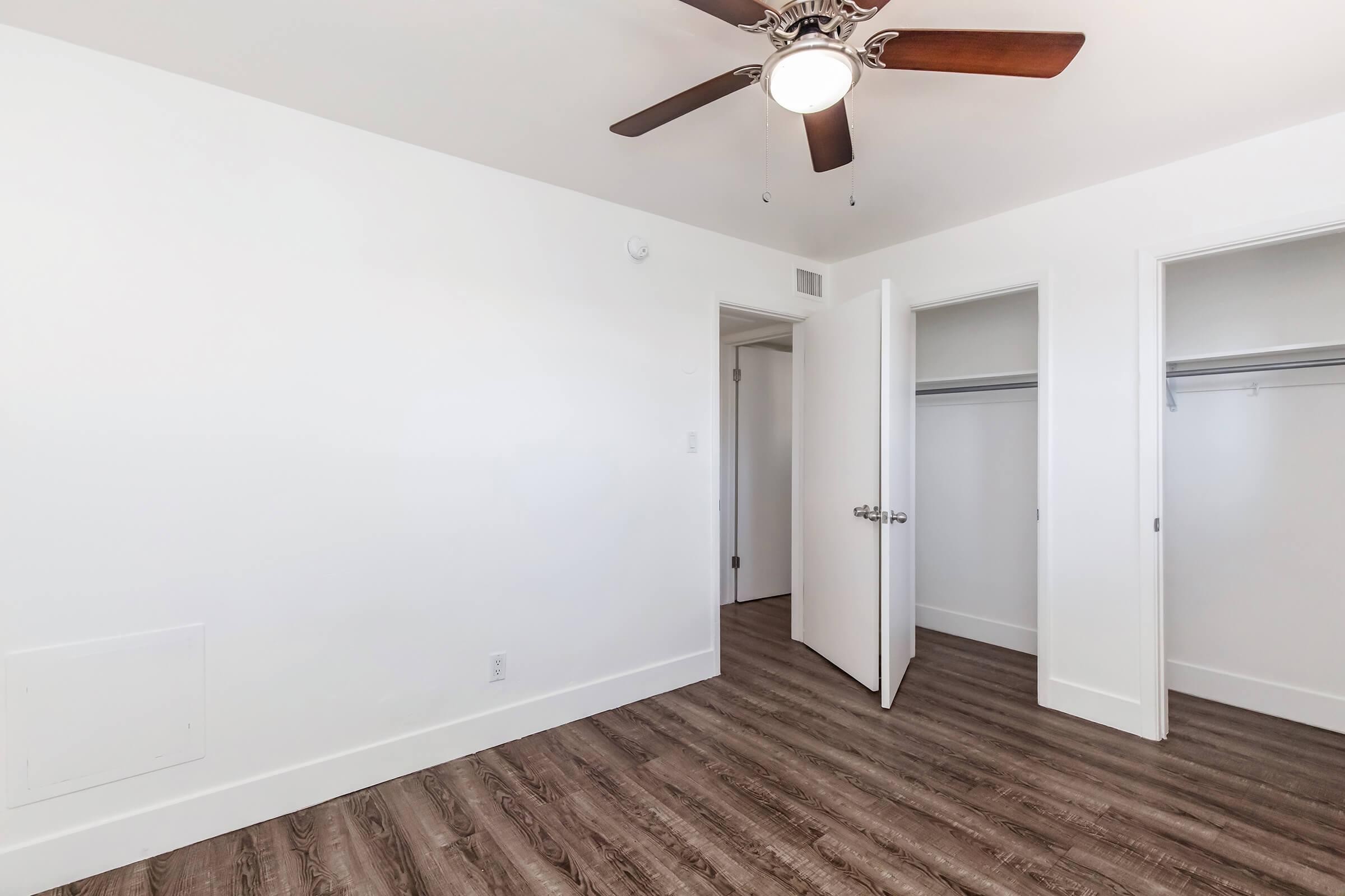 A clean, empty bedroom featuring light-colored walls, a ceiling fan with wooden blades, and two open closet doors. The floor is made of wood-like laminate, creating a bright and spacious atmosphere.