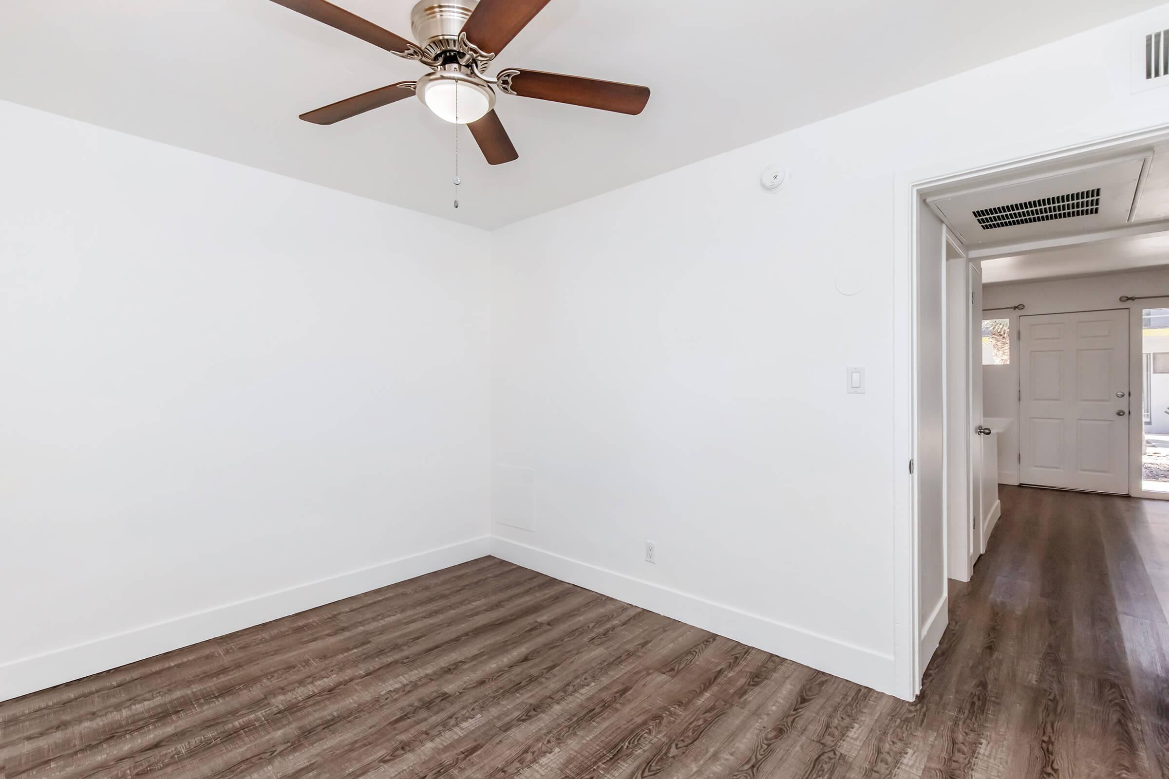 Empty room with a light-colored wall and a brown ceiling fan. The floor features dark wooden planks, and there is a door leading outside visible in the background. The room has no furniture, creating a spacious and minimalist atmosphere.