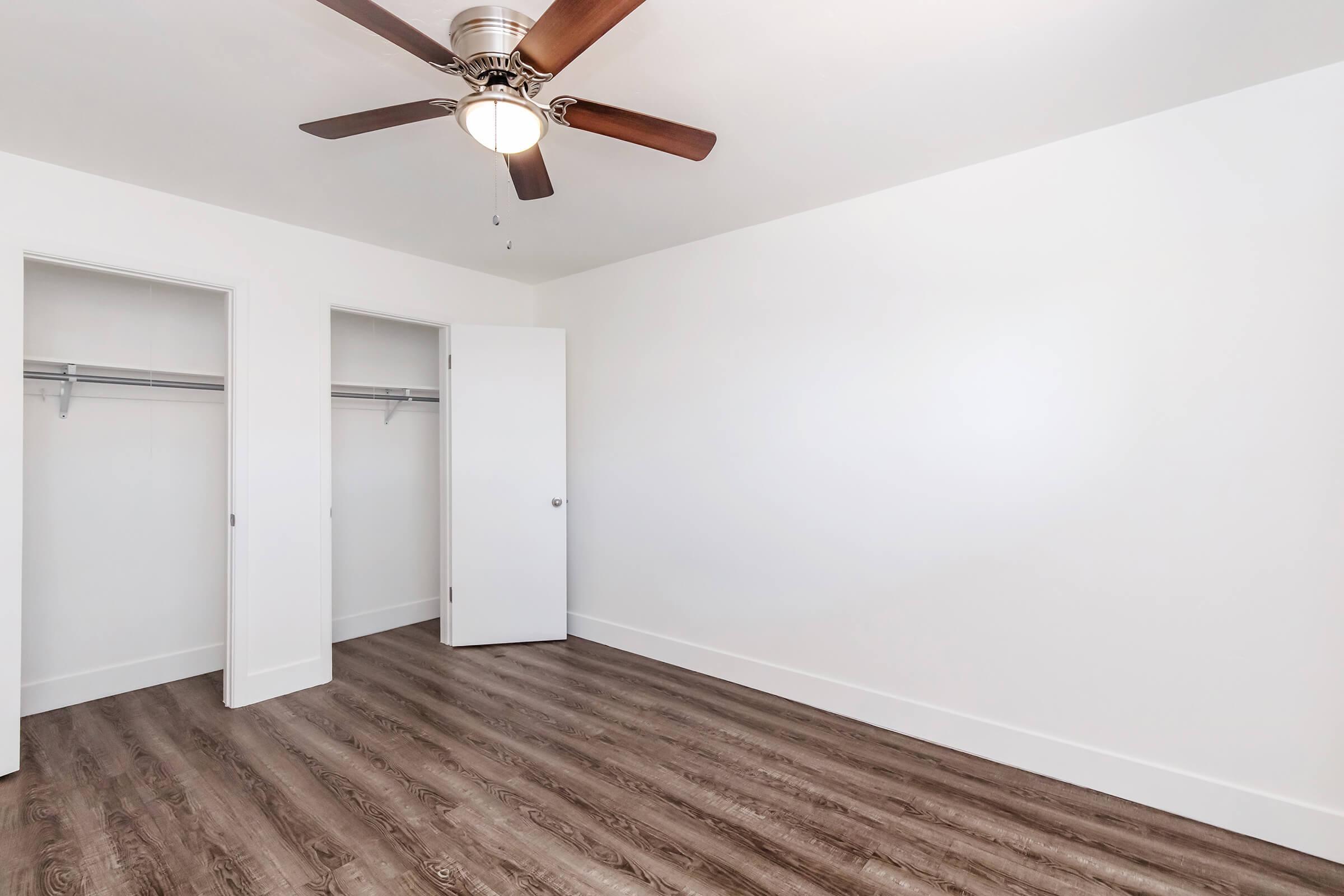 A bright, empty room featuring light-colored walls, a ceiling fan with wooden blades, and light brown wooden flooring. Two closets with open doors are visible on the left side. The space is uncluttered, giving a modern and airy feel.
