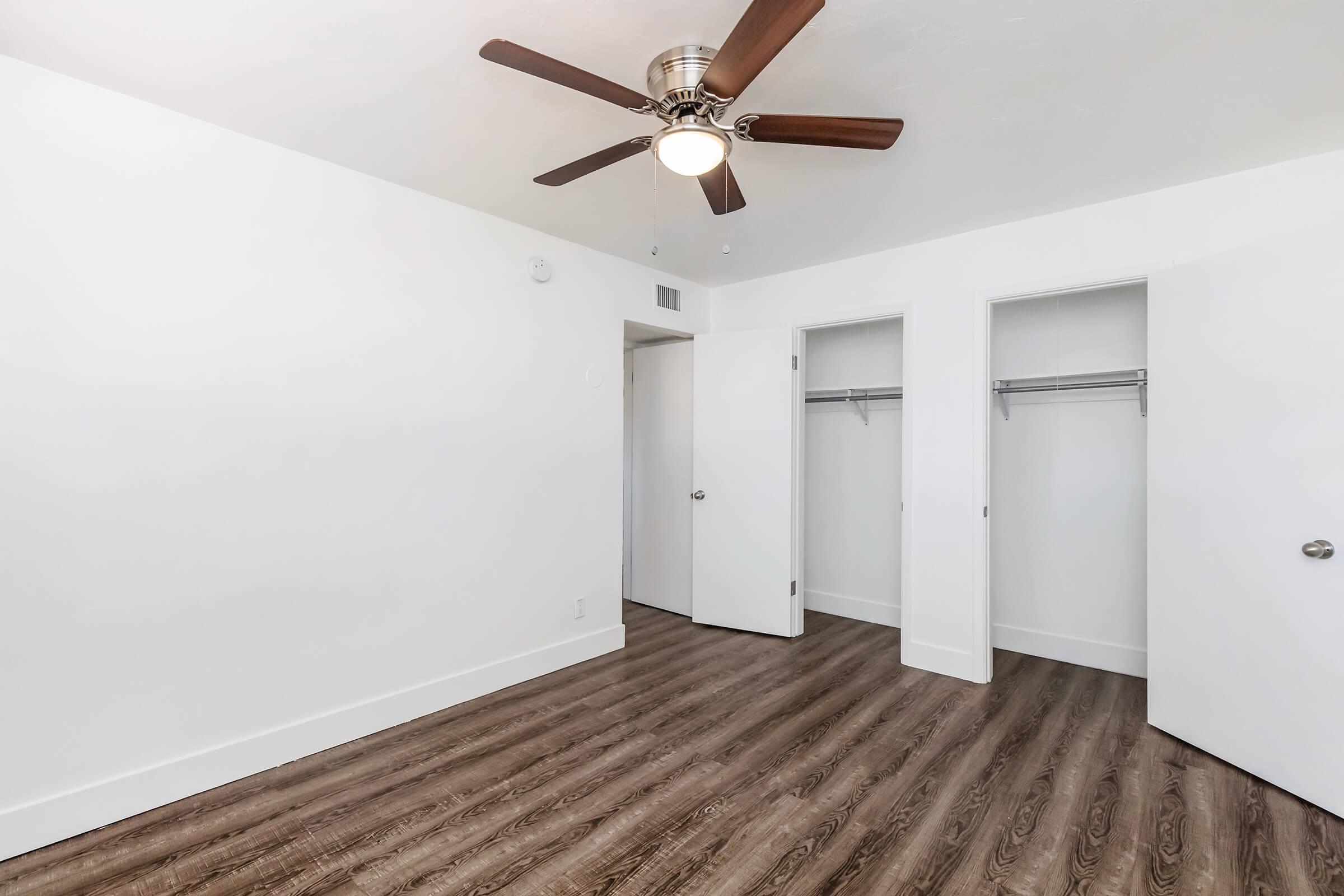 Bright, minimalistic room featuring a ceiling fan, light-colored walls, and a wood-look floor. Two open closet doors are visible, providing storage space. The overall design is modern and clean, creating a spacious and inviting atmosphere.