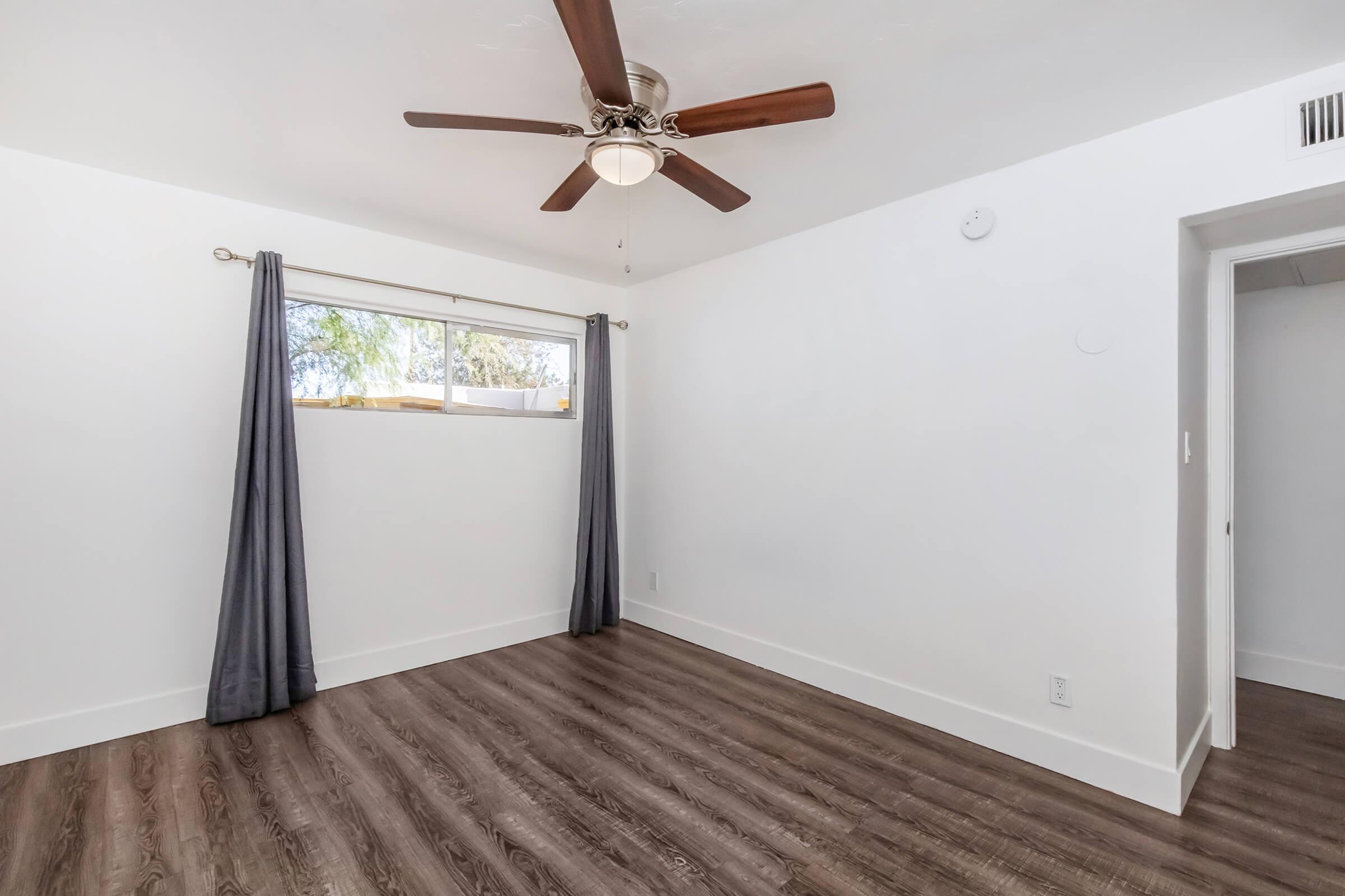 A bright, empty room featuring a ceiling fan with wooden blades, a window with sheer curtains allowing natural light, and light-colored walls. The floor is made of dark wood laminate, and there is a door on the right leading to another space.