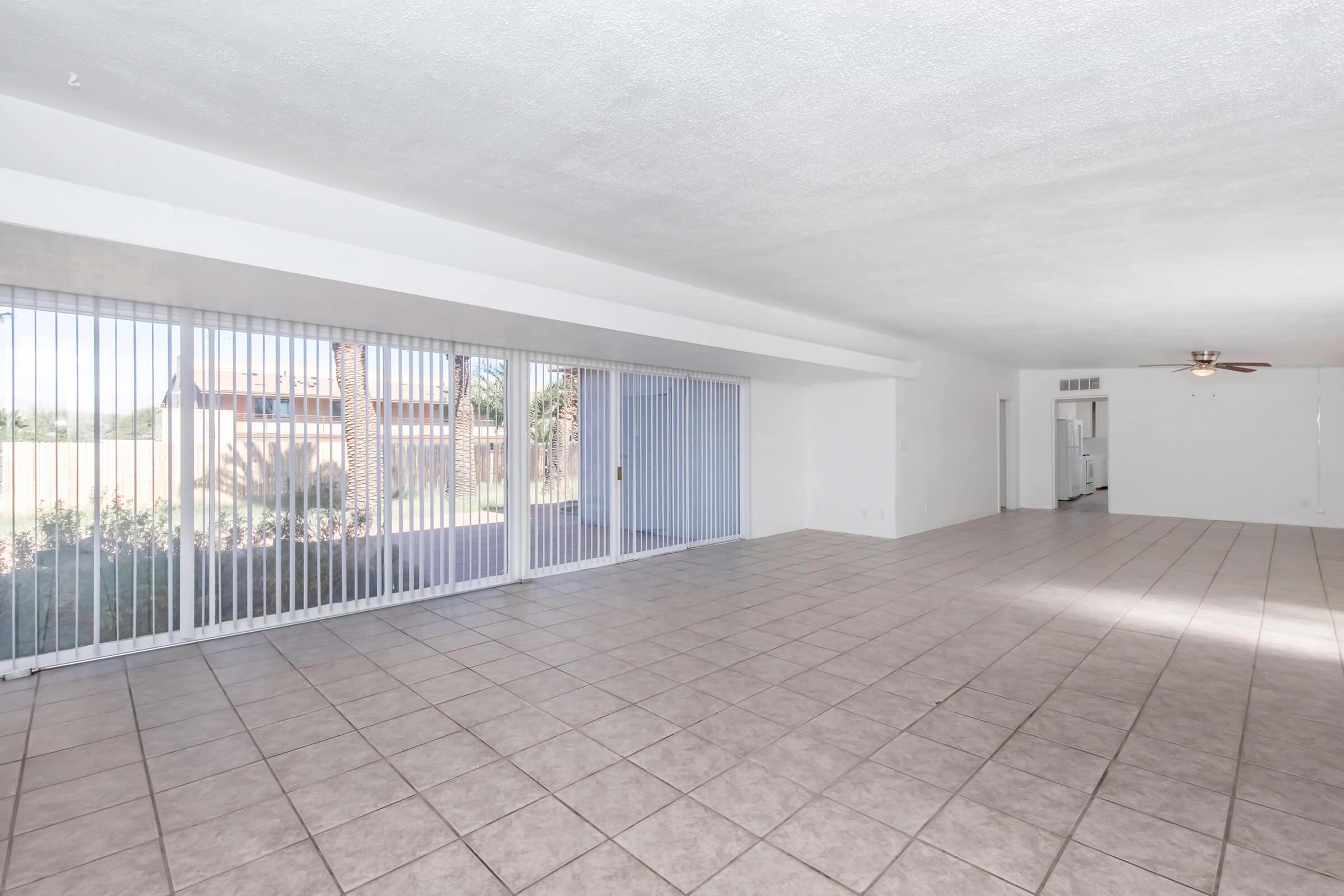 A spacious, open living area with tiled flooring, featuring large sliding glass doors that invite natural light. The room has a minimalistic design with white walls and a ceiling fan. A glimpse of an outdoor area with greenery can be seen through the glass doors.