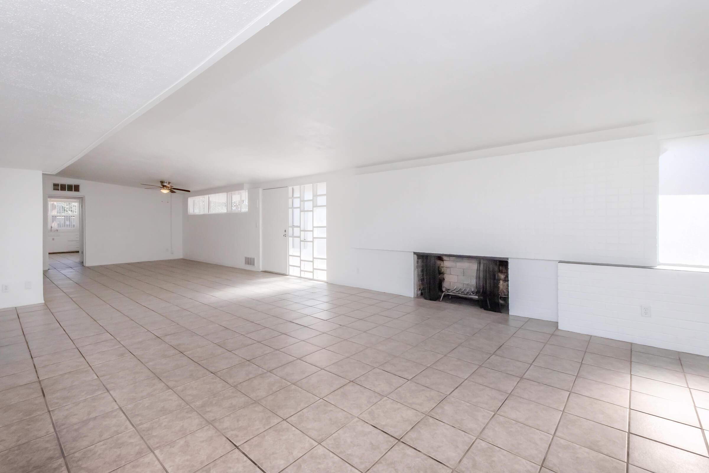 A spacious, empty living room with light-colored tile flooring, white walls, and a large window. There is a simple fireplace on one wall, and glass doors leading to another room or outside. The ceiling is plain and unobtrusive, adding to the bright and airy atmosphere of the space.