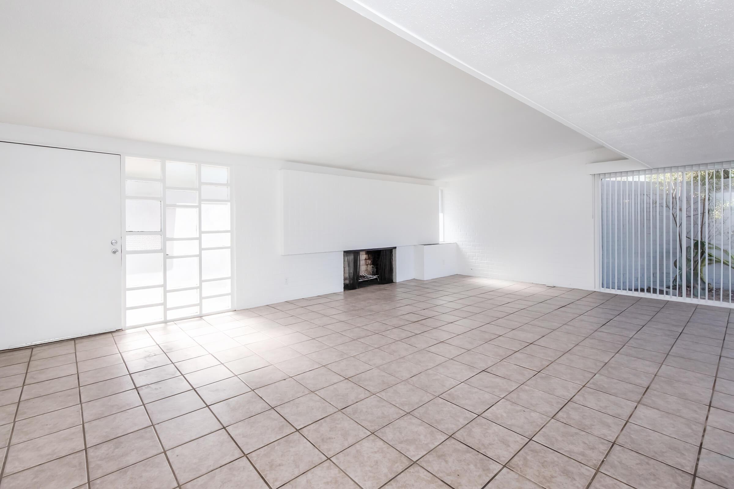 A spacious, empty room with light-colored walls and large windows. The floor is covered with square tiles, and there is a fireplace in the center of one wall. Natural light enters through the windows, enhancing the open and airy feel of the space.