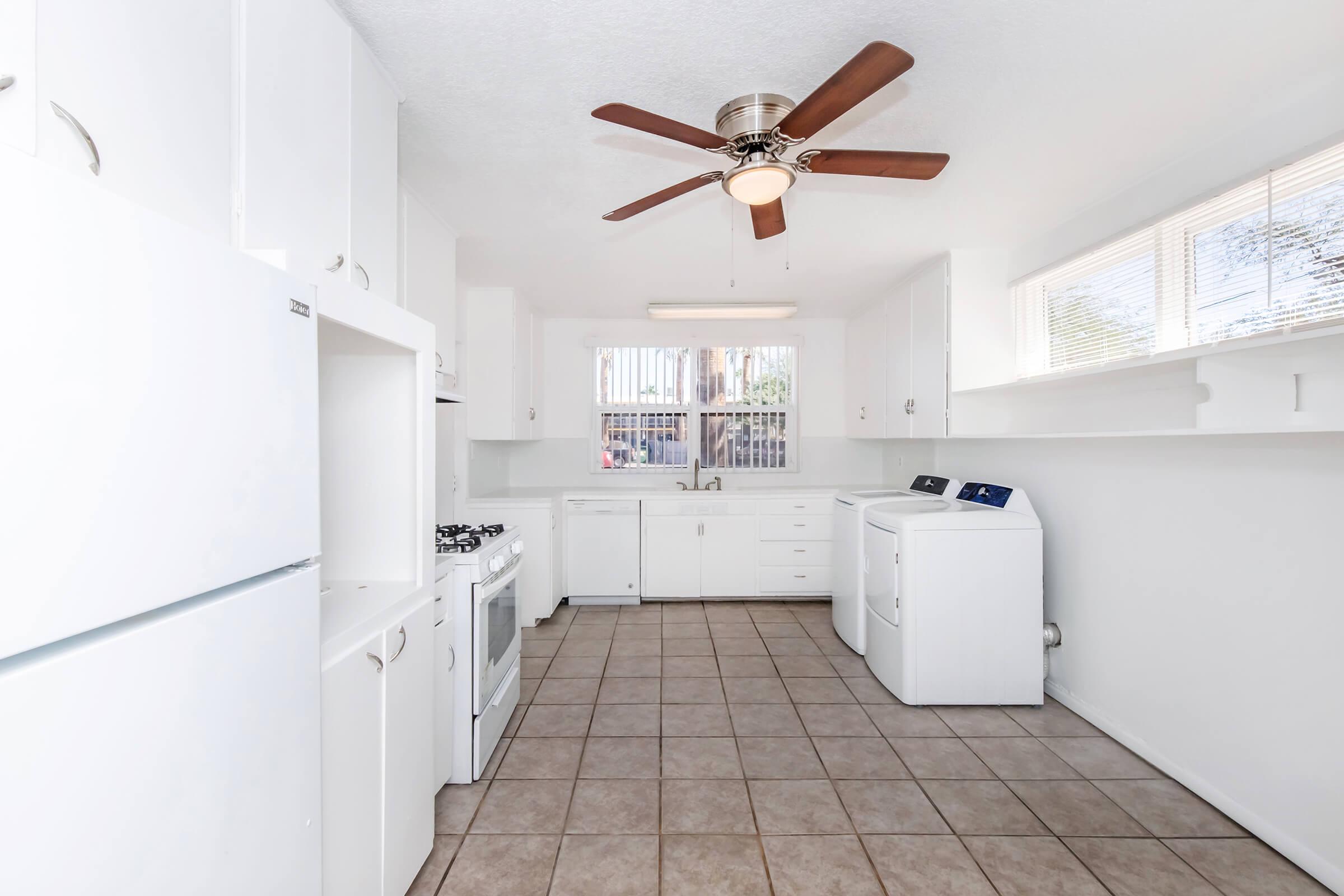 Bright and spacious kitchen featuring white appliances, a ceiling fan, and a window above the sink allowing natural light. The room includes a refrigerator, gas stove, and washer/dryer combo, with tiled flooring that enhances the clean, modern look.
