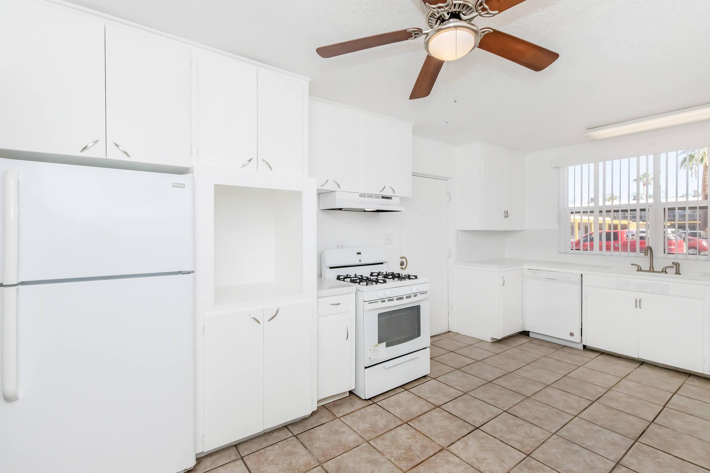 Bright and clean kitchen with white cabinets, a white refrigerator, and a white stove. Features a ceiling fan and large window with vertical blinds, allowing natural light. Tiled floor adds a contemporary touch.