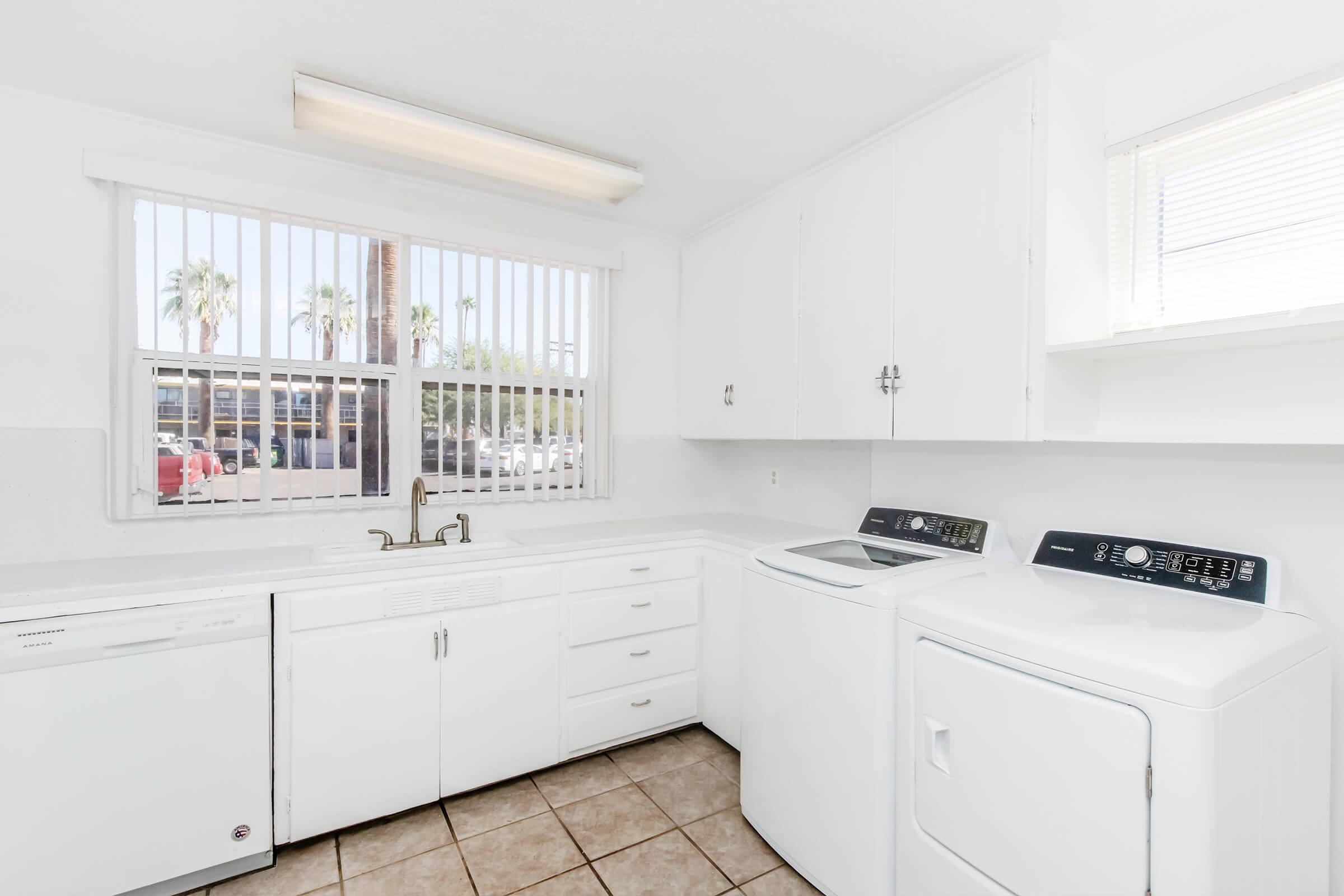Bright and clean laundry area featuring a white countertop, sink, dishwasher, and washer/dryer. Large window with vertical blinds allows natural light in, with a view of palm trees outside. The tiled floor adds a modern touch to the spacious layout.