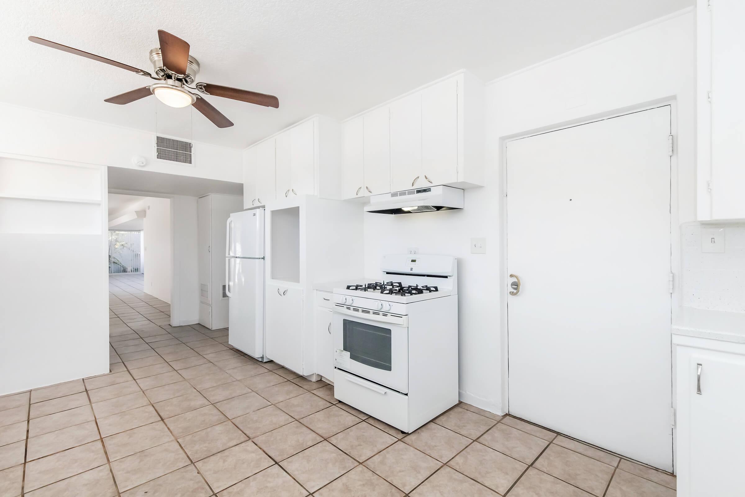 A bright, clean kitchen featuring white cabinets, a gas stove, and a refrigerator. The space has tiled flooring and a ceiling fan, with a door leading to another room. Natural light brightens the area, creating a welcoming atmosphere.