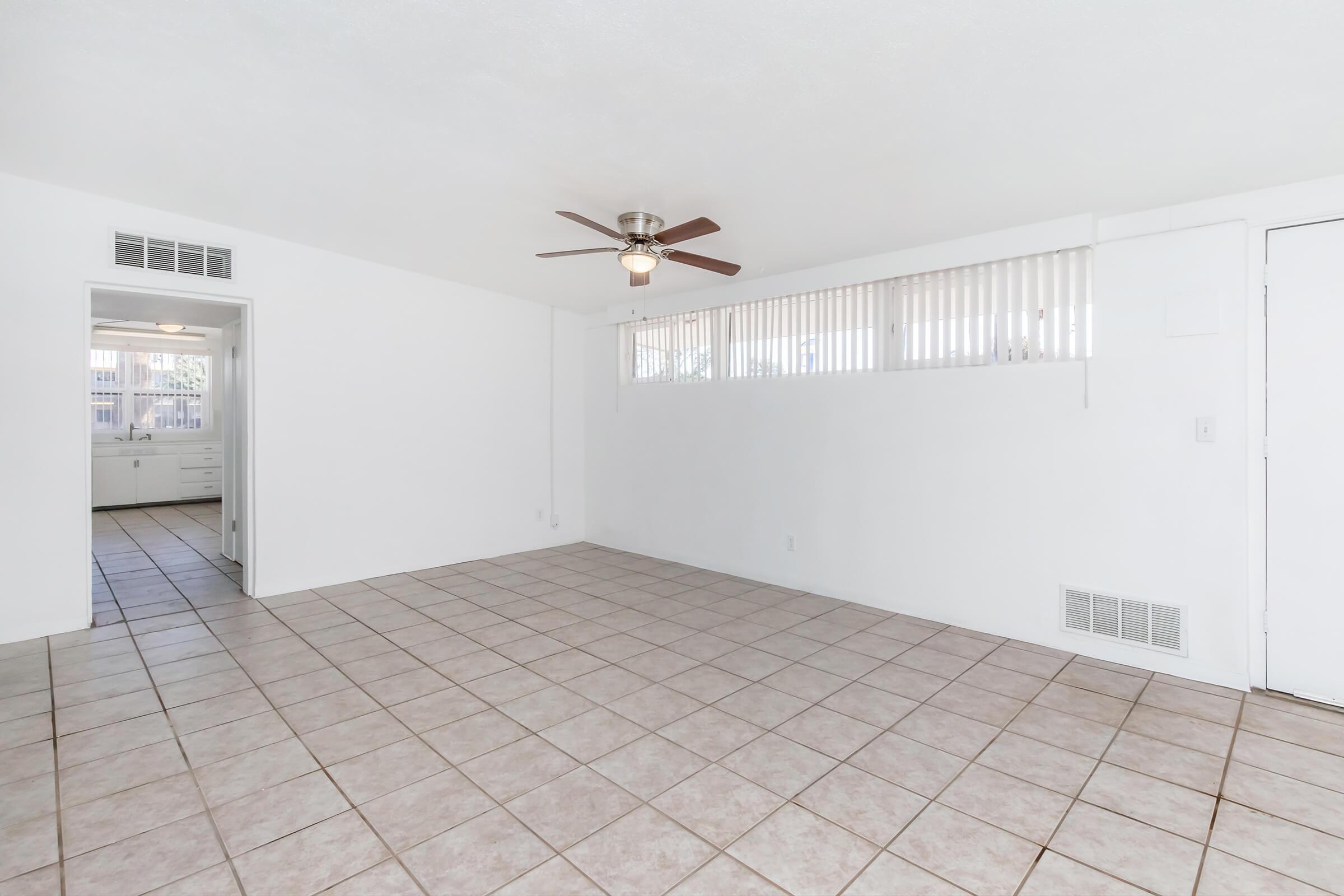 A spacious, empty room with white walls and tiled flooring. There is a ceiling fan in the center, and a wall of windows with blinds on one side. An entrance to a kitchen is visible in the background. The overall atmosphere is bright and airy, suitable for various setups or furnishings.