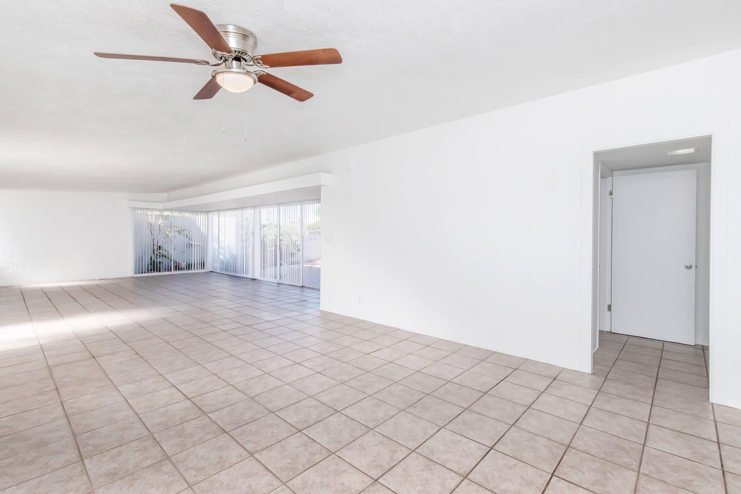 A spacious, empty living area with light-colored walls and tiled flooring. It features a ceiling fan and large windows covered with sheer curtains, allowing natural light to enter. A door on the right leads to another room or hallway. The overall ambiance is bright and airy.