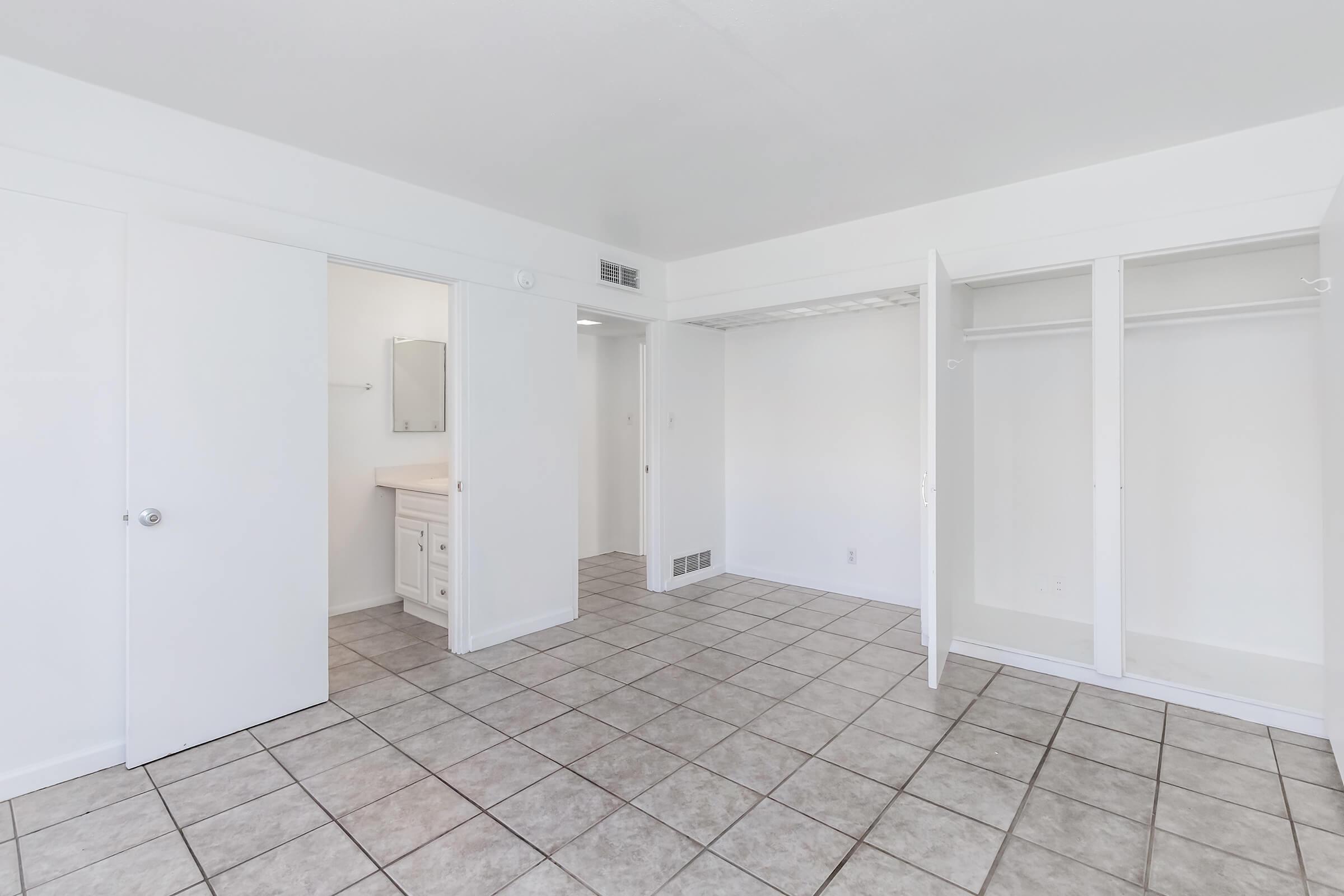 A bright, empty room with white walls and tiled flooring. On one side, there are two open closet spaces, and a bathroom area with a mirror visible. The room has a clean, minimalist aesthetic, featuring several doors leading to other spaces. Natural light illuminates the area, creating an inviting atmosphere.