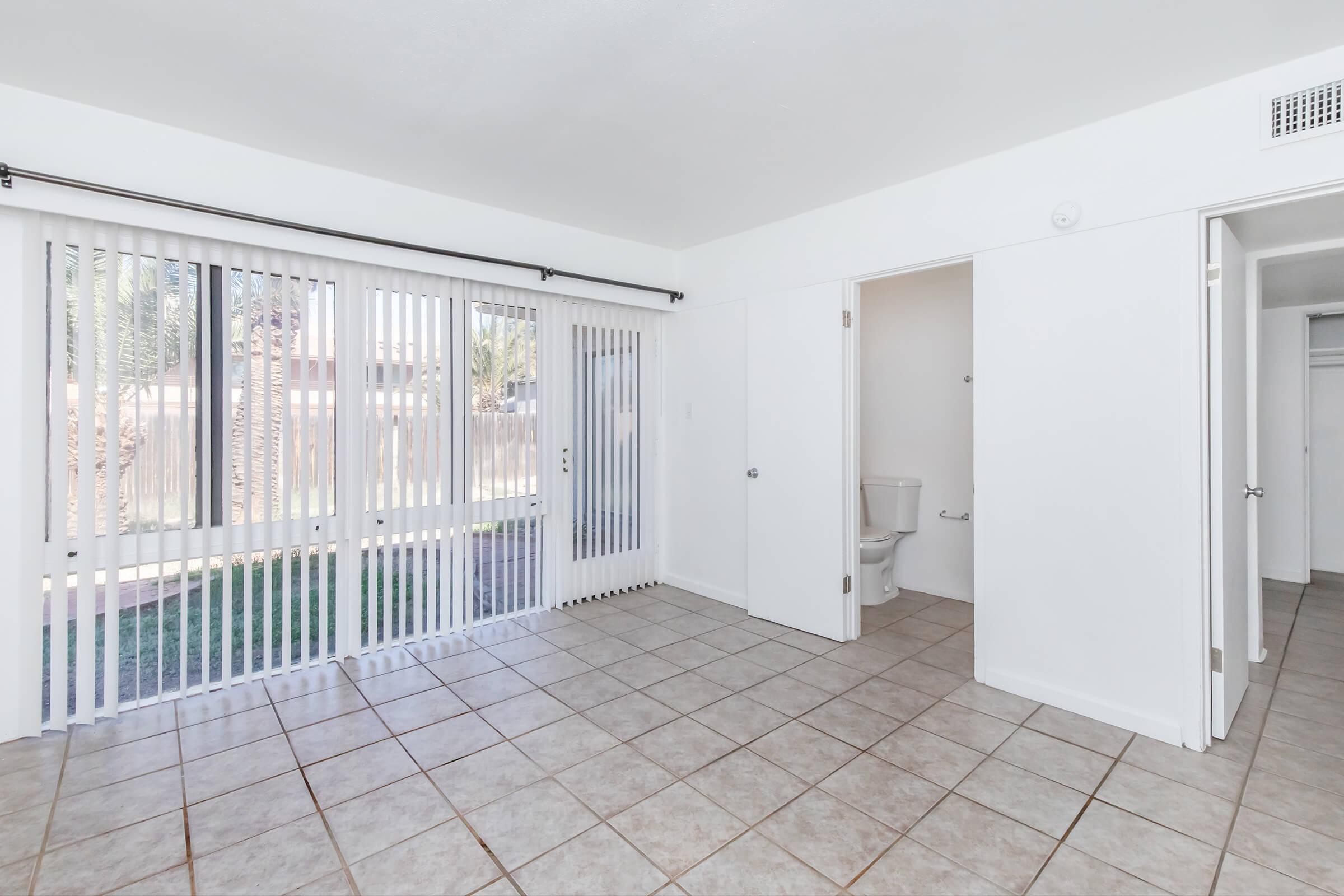 Spacious empty room featuring tile flooring, large sliding glass doors with vertical blinds, and a doorway leading to a bathroom. The walls are painted white, and there is a closet opening visible on the right side. Natural light enters through the doors, illuminating the space.