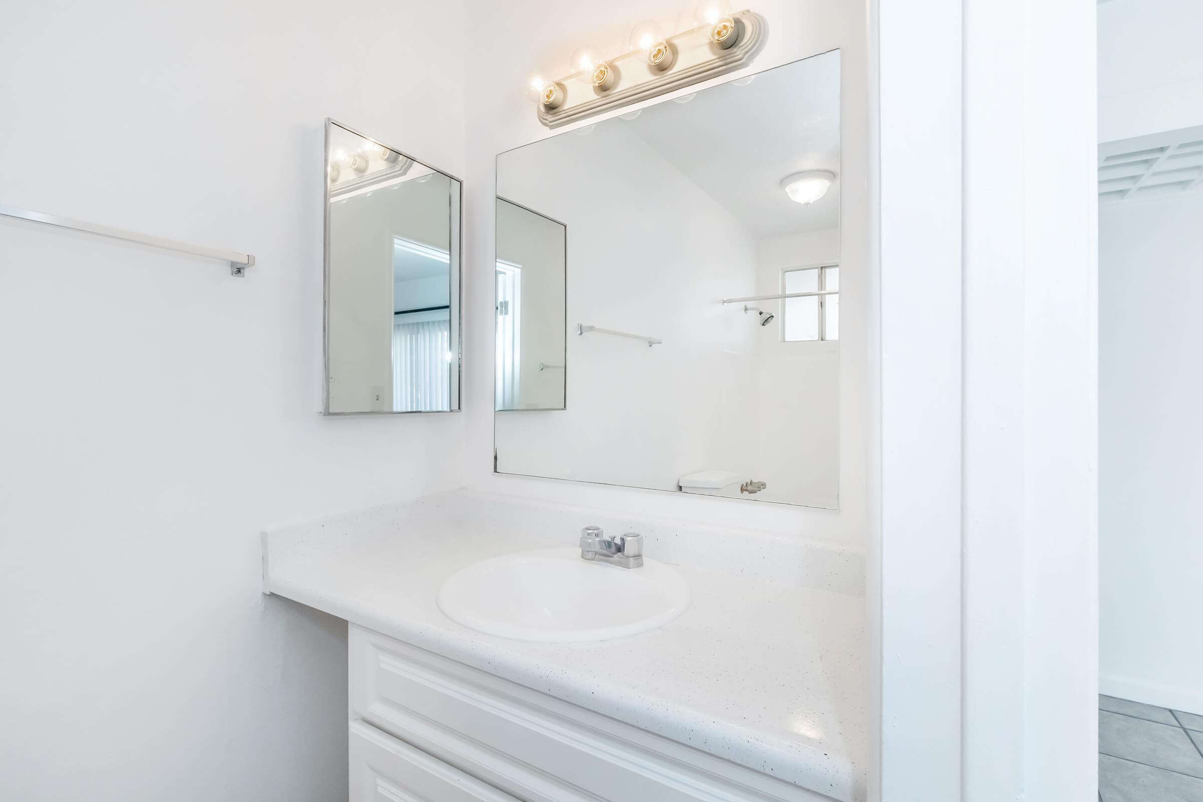 A clean, modern bathroom featuring a white countertop with a round sink, a mirror above, and a simple light fixture. The walls are painted white, and there is a towel rack on the left. A shower area is visible in the background, contributing to the overall spacious feel of the room.