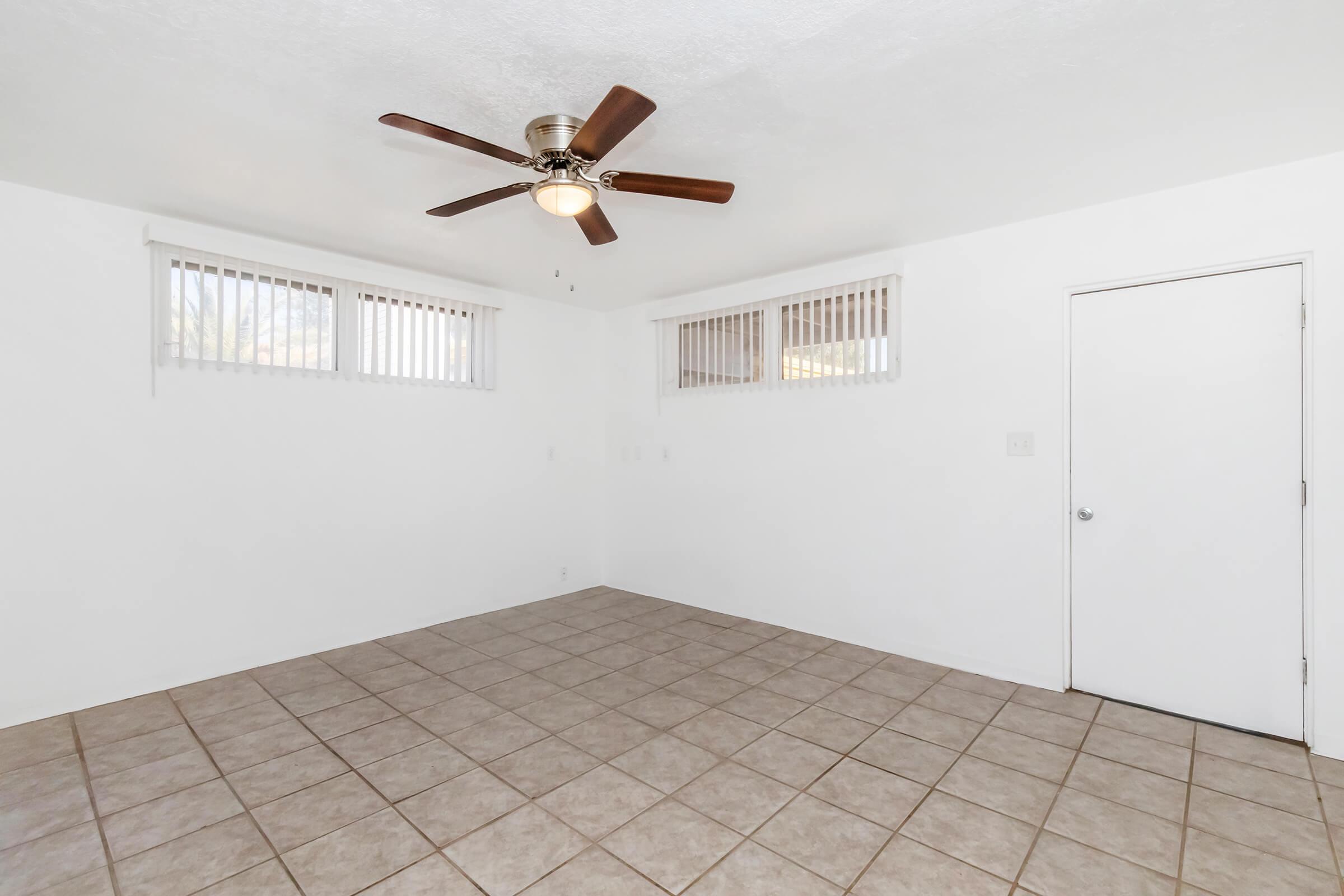 Interior of a room with white walls and tiled flooring. It features a ceiling fan with wooden blades and two small windows with horizontal blinds. A door is visible on the right side, leading to another space. The room appears empty and well-lit, creating a clean and spacious feel.