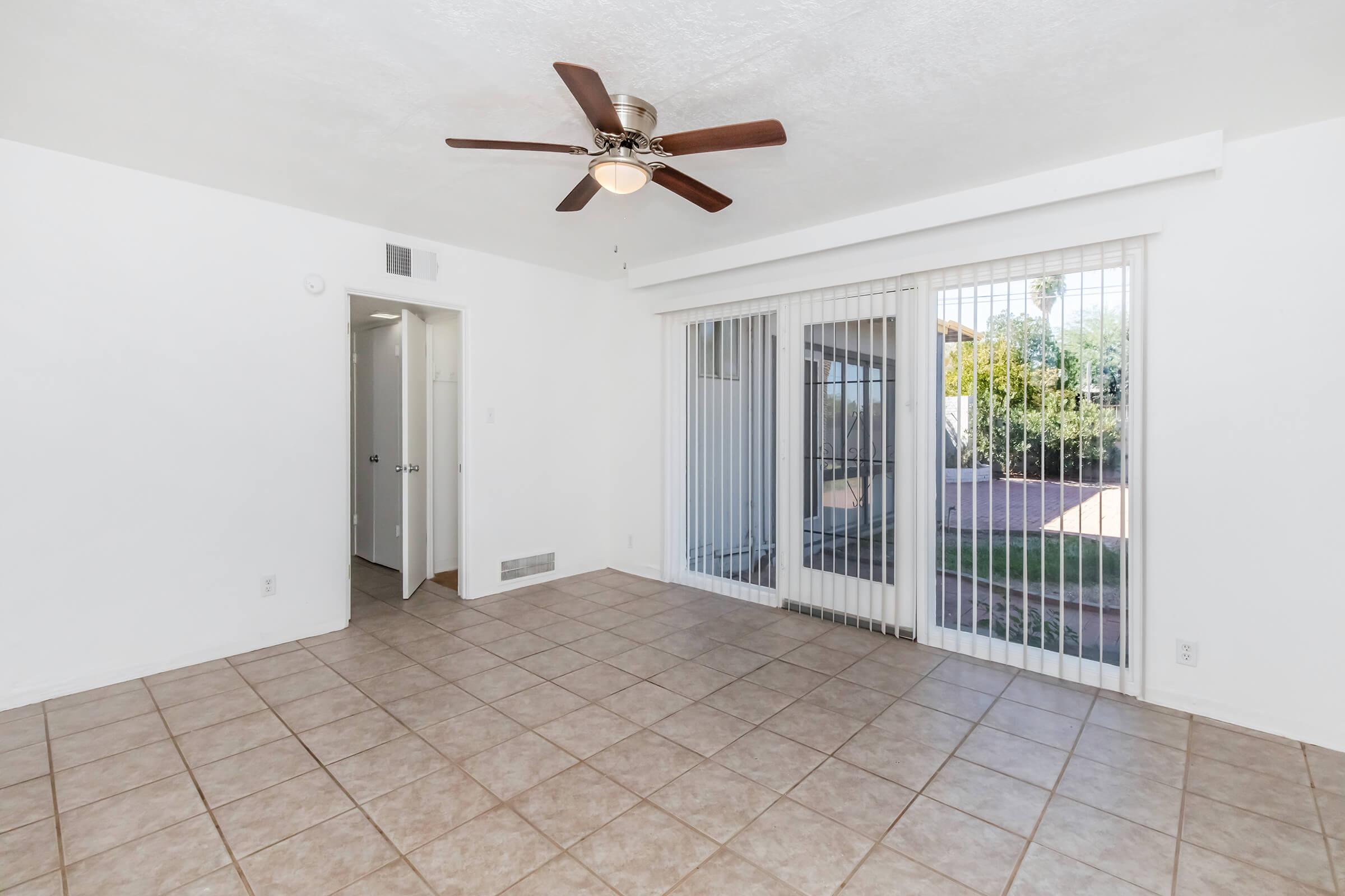 Spacious, well-lit living area with tile flooring, a ceiling fan, and sliding glass doors leading to an outdoor space. The room features white walls and a door that opens to a hallway, creating a fresh and inviting ambiance.