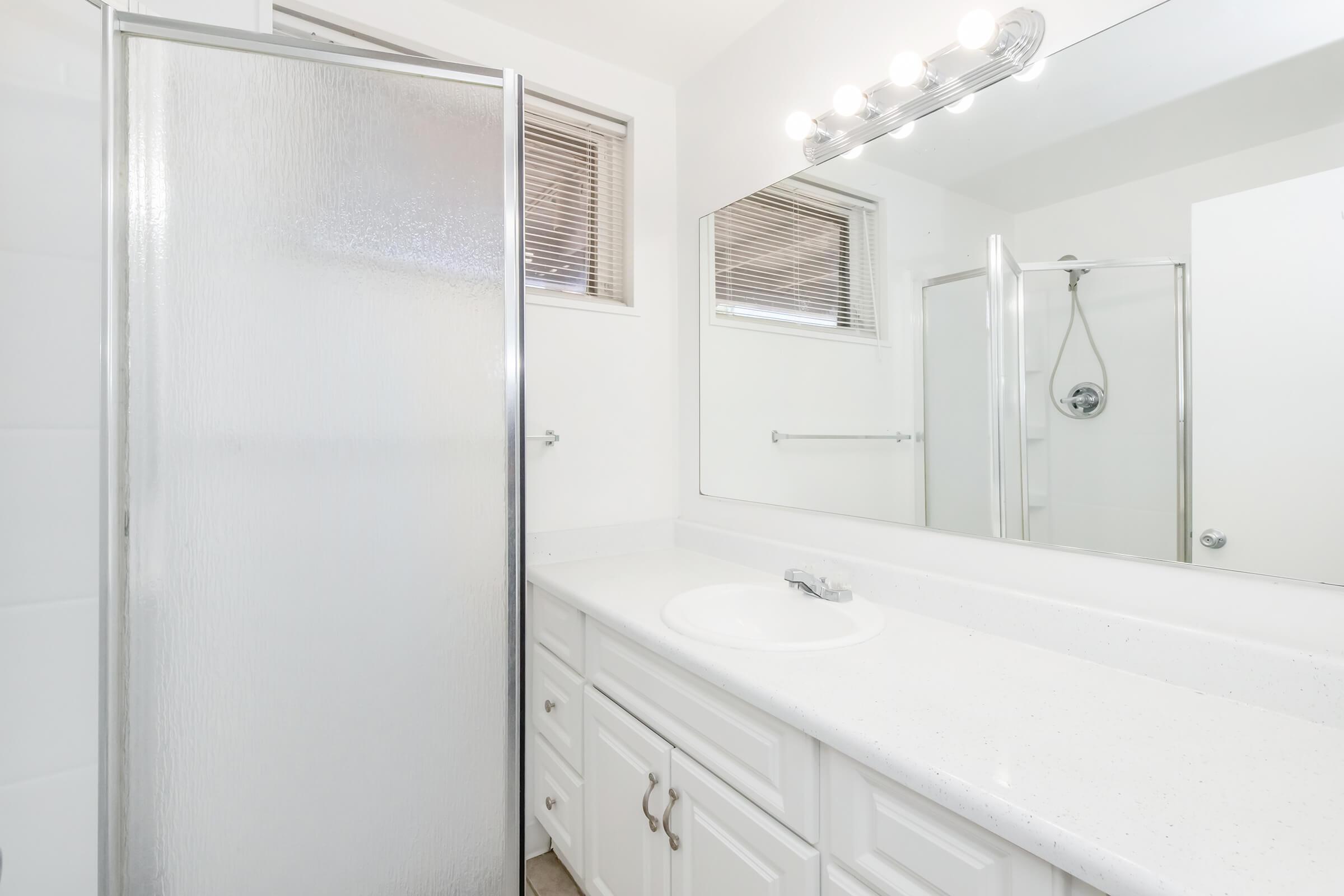 A clean, well-lit bathroom featuring a white countertop with a sink, a large mirror with overhead lighting, a glass shower enclosure, and a small window. The walls are painted in a light color, and the floor is tiled, creating a modern and spacious atmosphere.