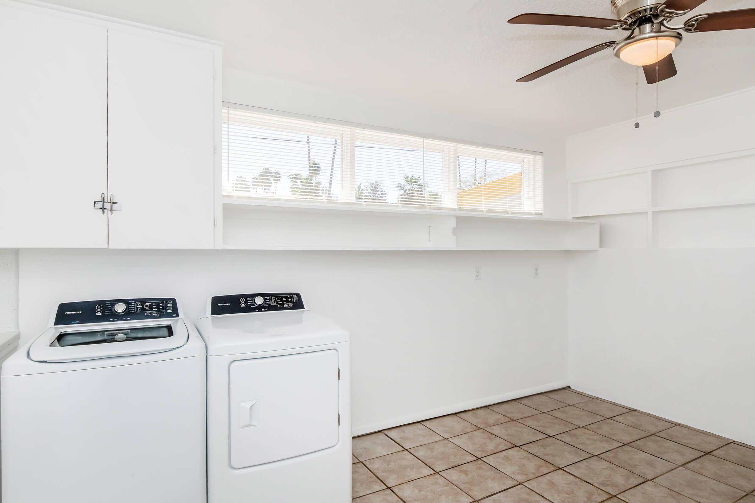 A bright laundry room featuring a washer and dryer, white cabinetry, and a ceiling fan. Large windows provide natural light, and there are shelves along the wall for additional storage. The floor is tiled, creating a clean and functional space.