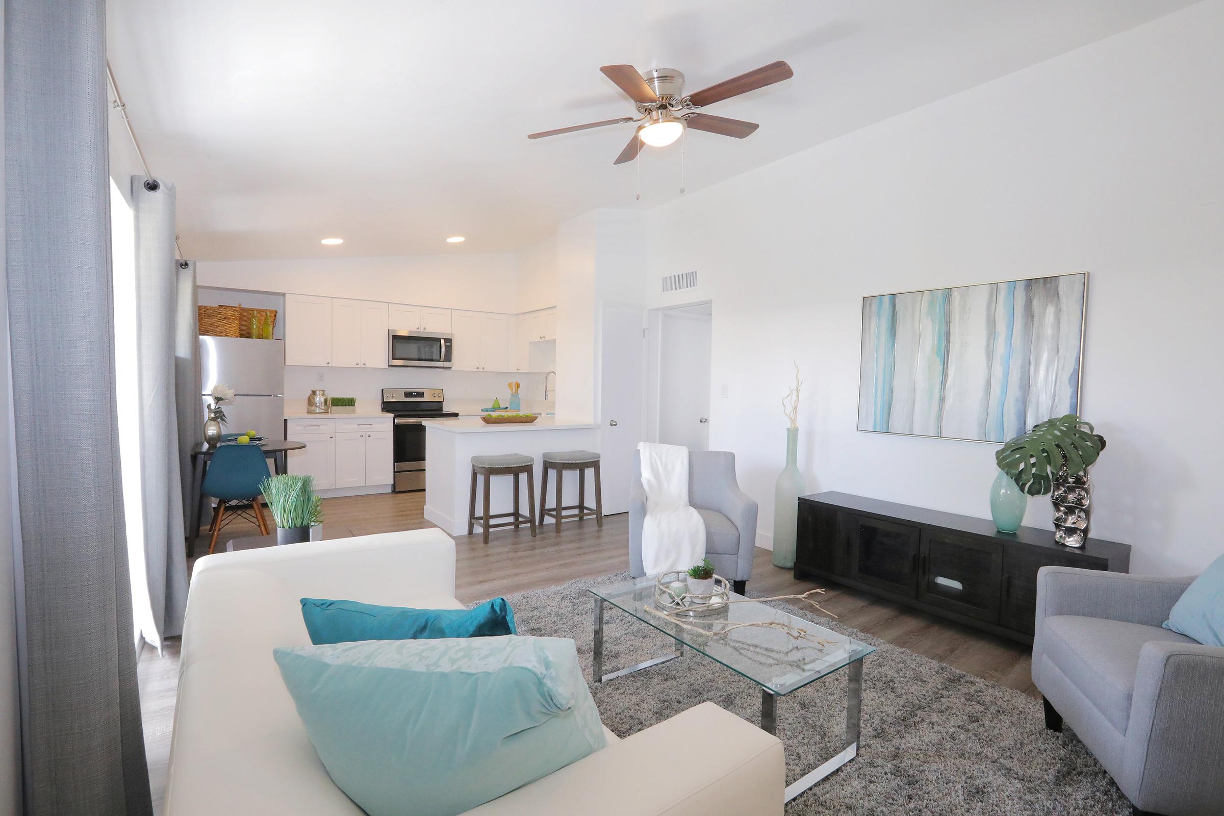 A bright and modern living room featuring a white sofa with teal cushions, a glass coffee table, and a dark wooden TV stand. There is a ceiling fan above, a colorful abstract painting on the wall, and a view of a well-equipped kitchen with light cabinets in the background. Natural light fills the space through large windows.