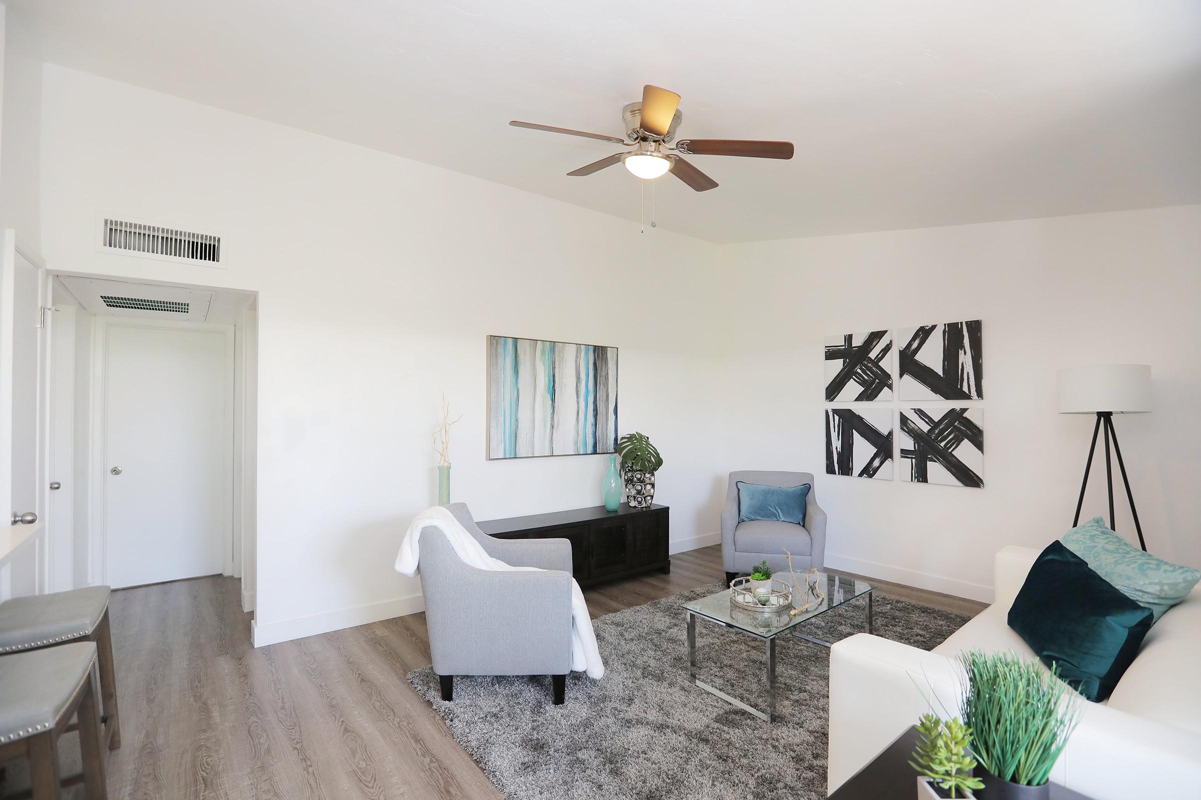 A modern living room featuring light-colored walls, a ceiling fan, and stylish furnishings. There are two gray armchairs, a glass coffee table with decorative items, and a black console table. Artwork hangs on the wall, and a floor lamp adds ambient lighting, creating a cozy and inviting atmosphere.