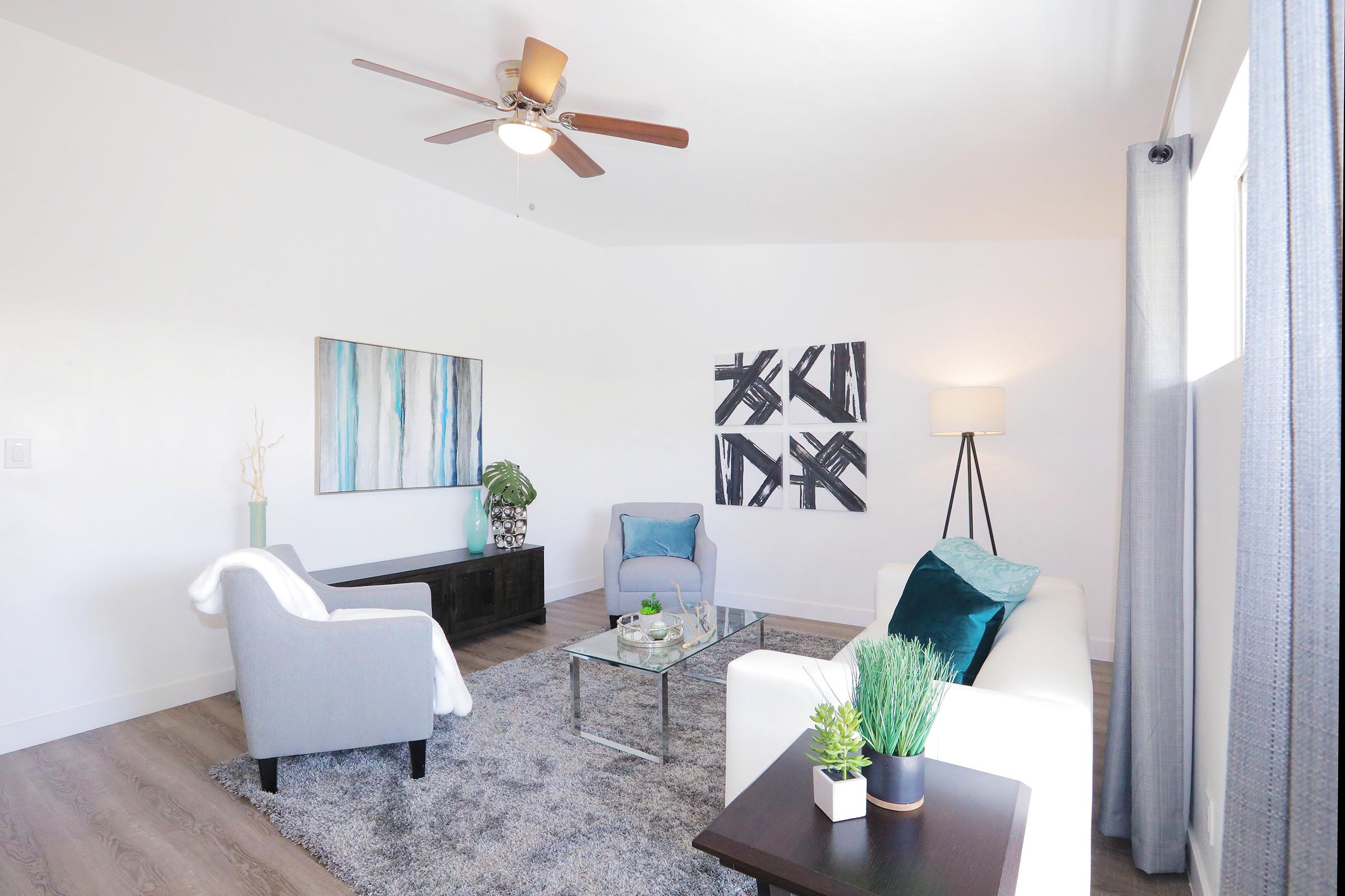A bright, modern living room featuring a white sofa with teal cushions, a gray armchair, and a dark wood console table. The space includes a gray area rug, potted plants, wall art with abstract designs, and a standing lamp, all under a ceiling fan. Natural light streams in through a window with sheer curtains.