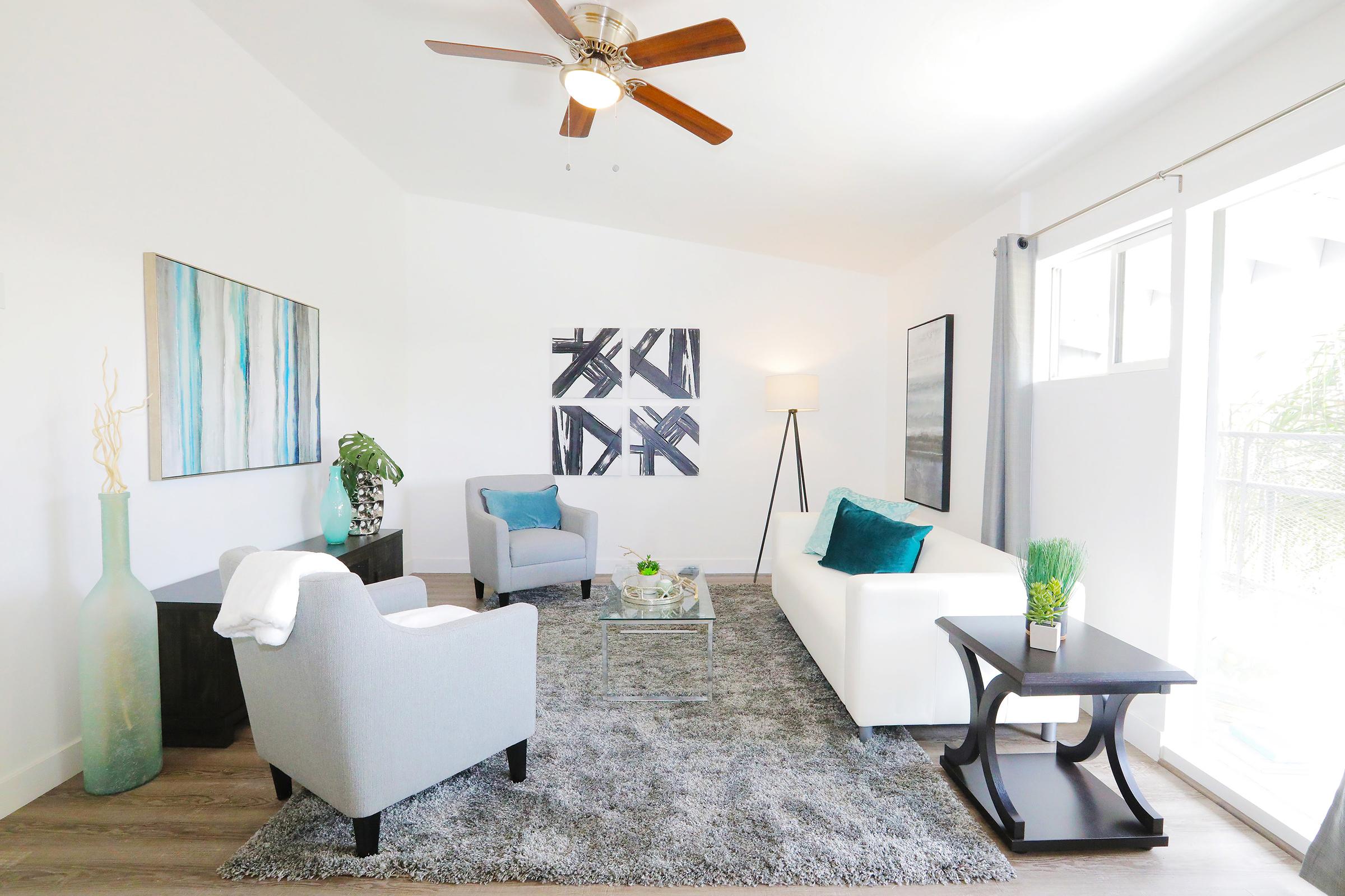 Bright and modern living room featuring a white sofa, gray armchairs, and a stylish coffee table. Decor includes abstract wall art, a potted plant, and a light fixture. A large window allows natural light to fill the space, which has neutral-colored walls and a plush gray rug.
