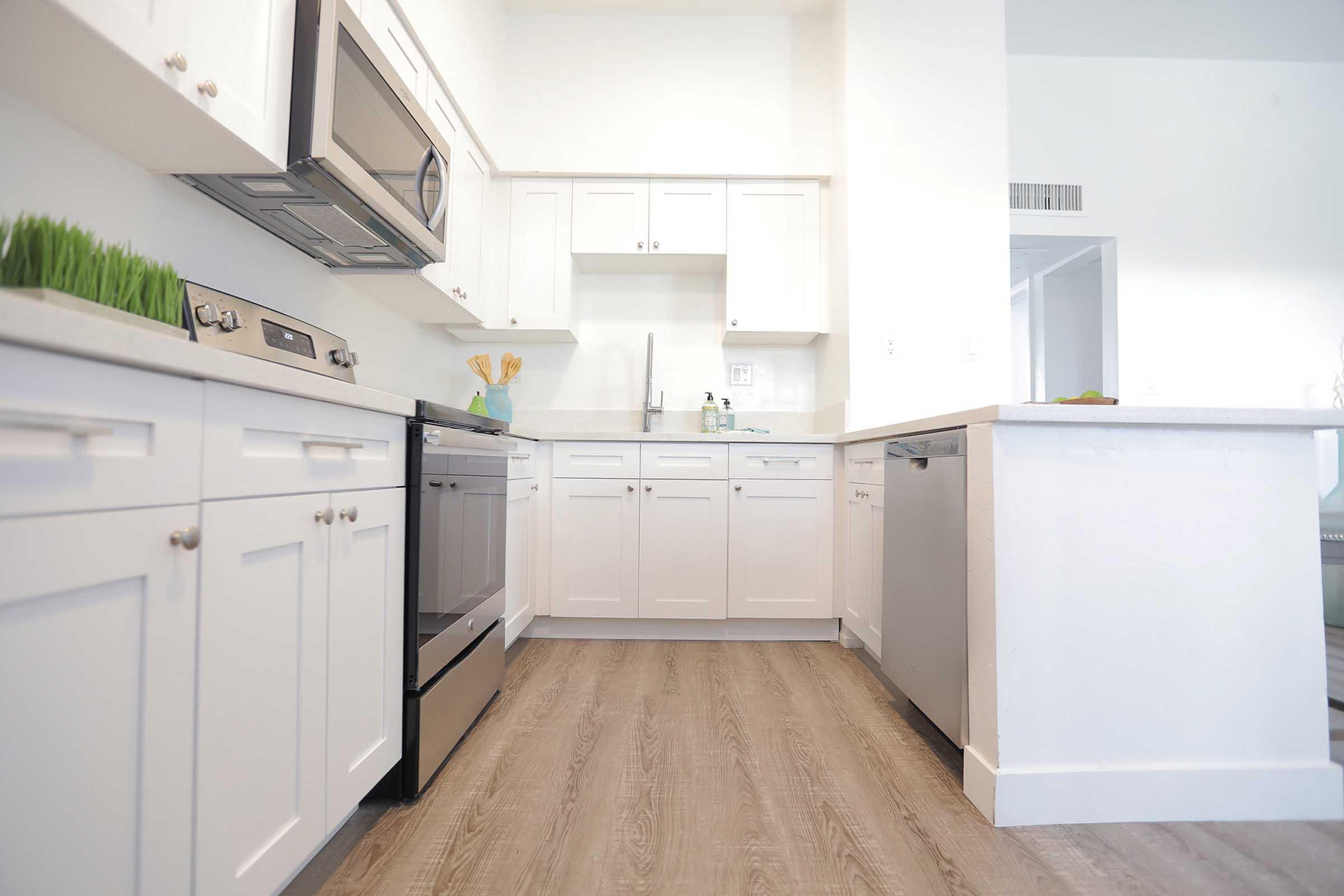Modern kitchen featuring white cabinets, stainless steel appliances, and a seamless counter. Natural light fills the space, highlighting the wood-like flooring and simple, elegant design. A small green plant adds a touch of color to the countertop, creating a fresh and inviting atmosphere.