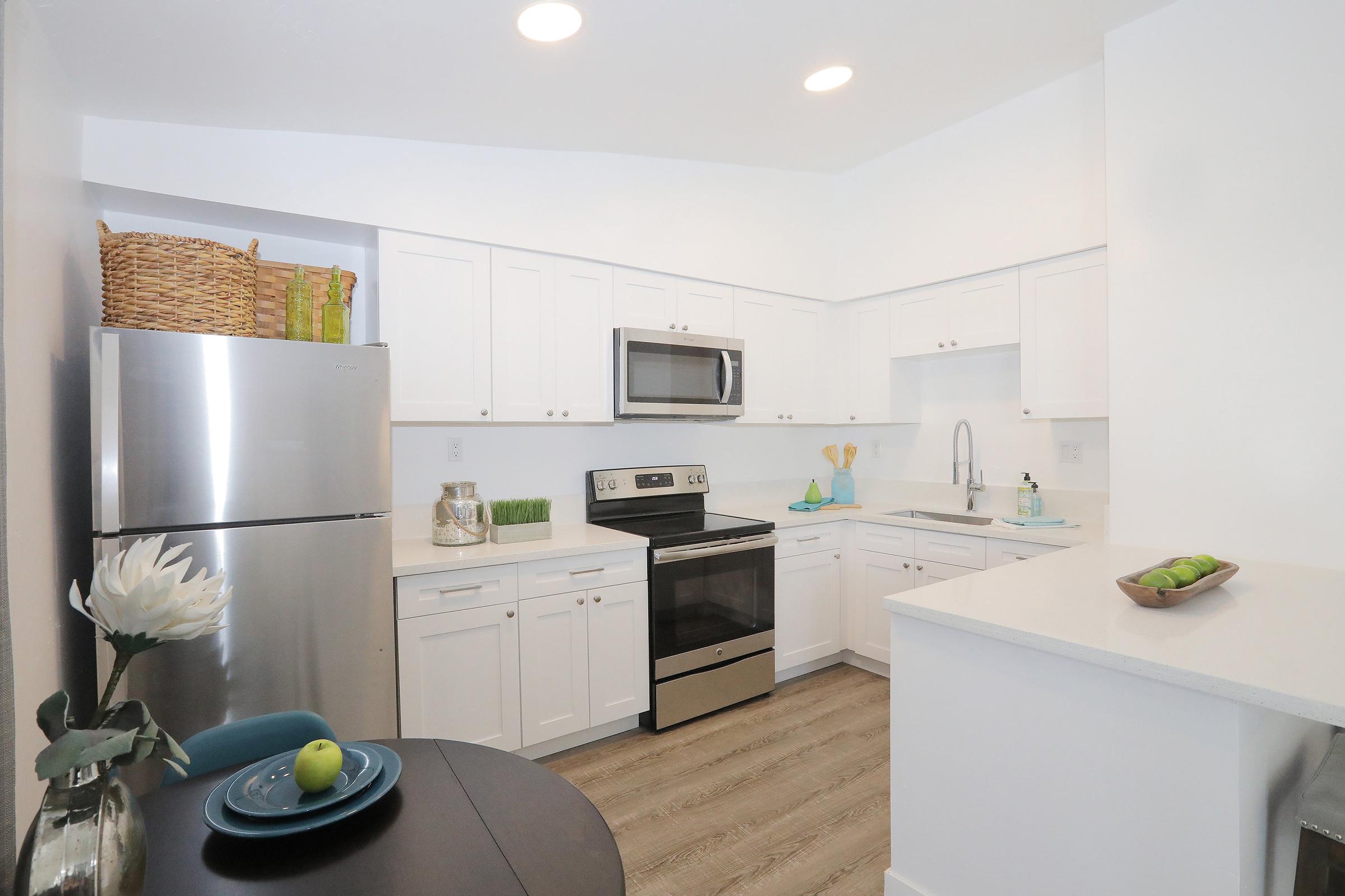 Modern kitchen featuring white cabinetry, stainless steel appliances, and a clean countertop. A sleek refrigerator is on the left, with a microwave and oven built into the cabinetry. Minimalist décor includes a small table set with a plate and a decorative bowl, along with a basket on top of the cabinets.