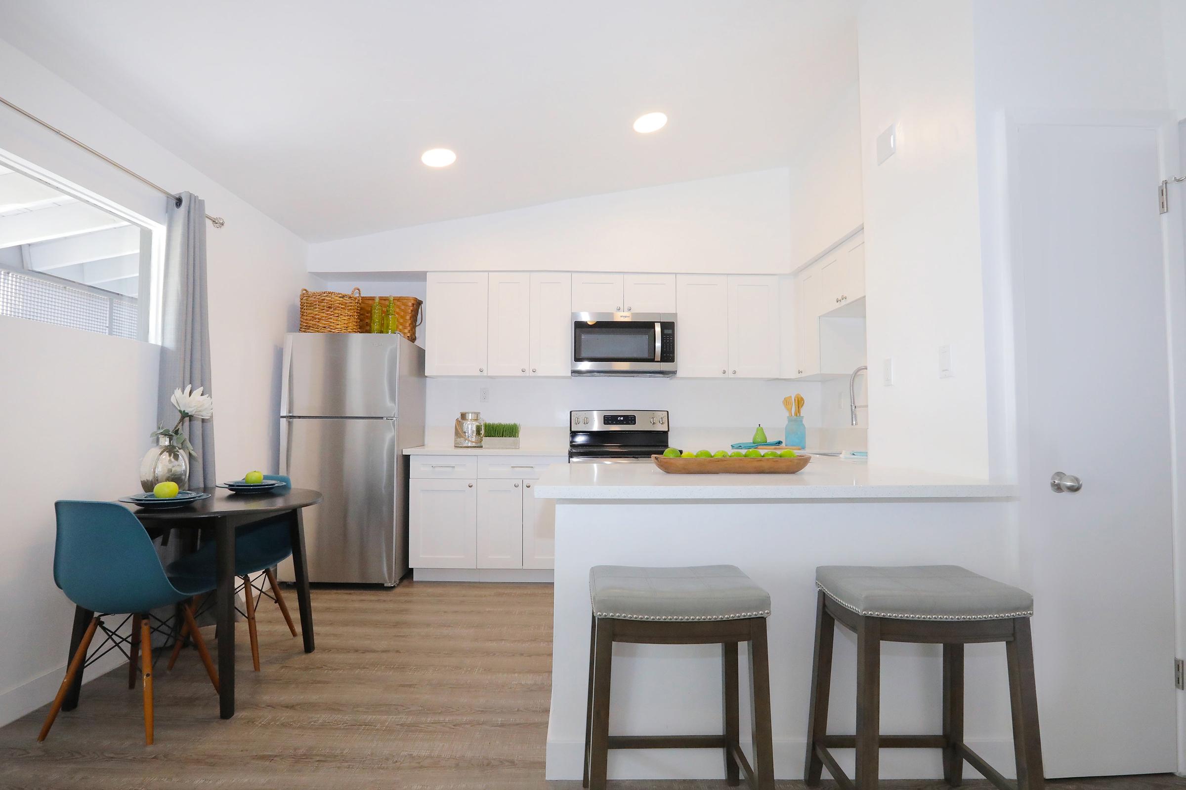 Modern kitchen featuring white cabinetry, stainless steel appliances, and a white countertop. A small dining table with blue chairs is set for two, and there are decorative items like a bowl of limes on the counter. The space is bright with natural light coming through a window.