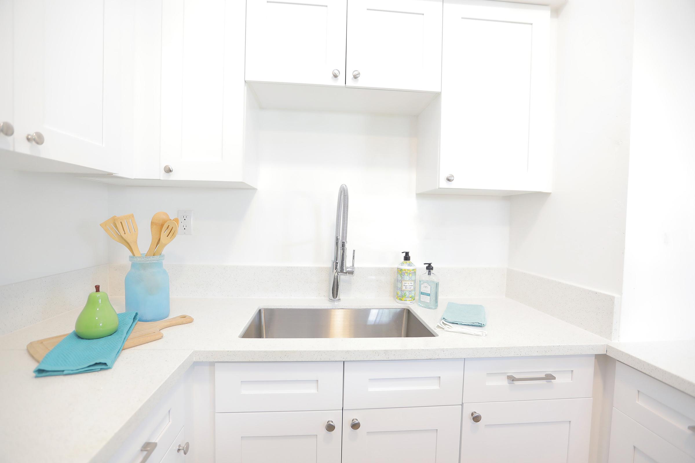 A modern kitchen sink area featuring white cabinetry, a stainless steel faucet, and a clean countertop. Kitchen utensils are displayed in a glass jar, a green fruit sits beside a wooden cutting board, and dish soap is neatly arranged nearby, creating a tidy and contemporary look.
