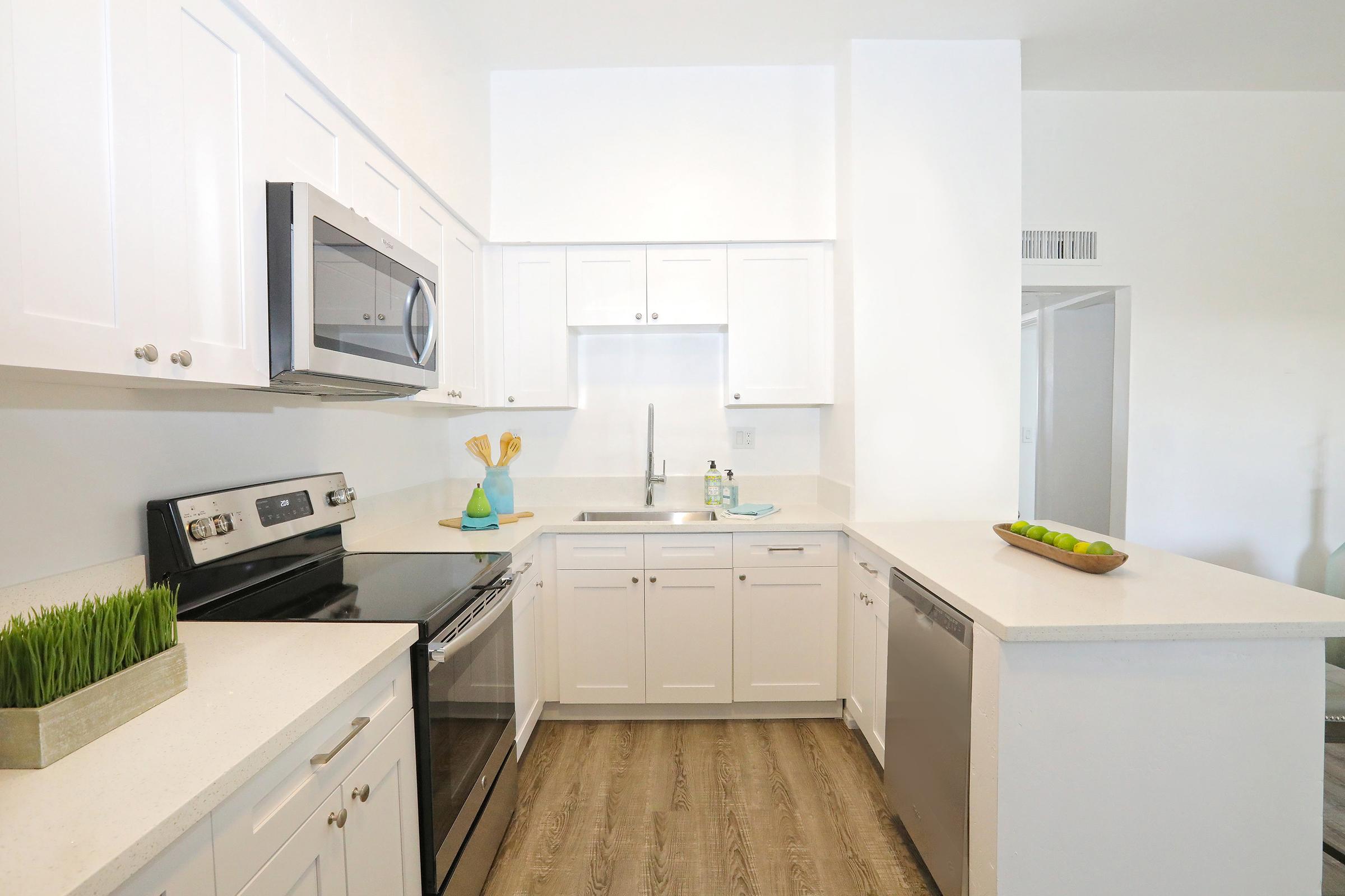 Modern kitchen featuring stainless steel appliances, white cabinetry, and a large countertop. There's a decorative bowl with green apples, a vase with wheat, and a small plant. The kitchen has a clean and spacious layout, with light-colored walls and wooden flooring.