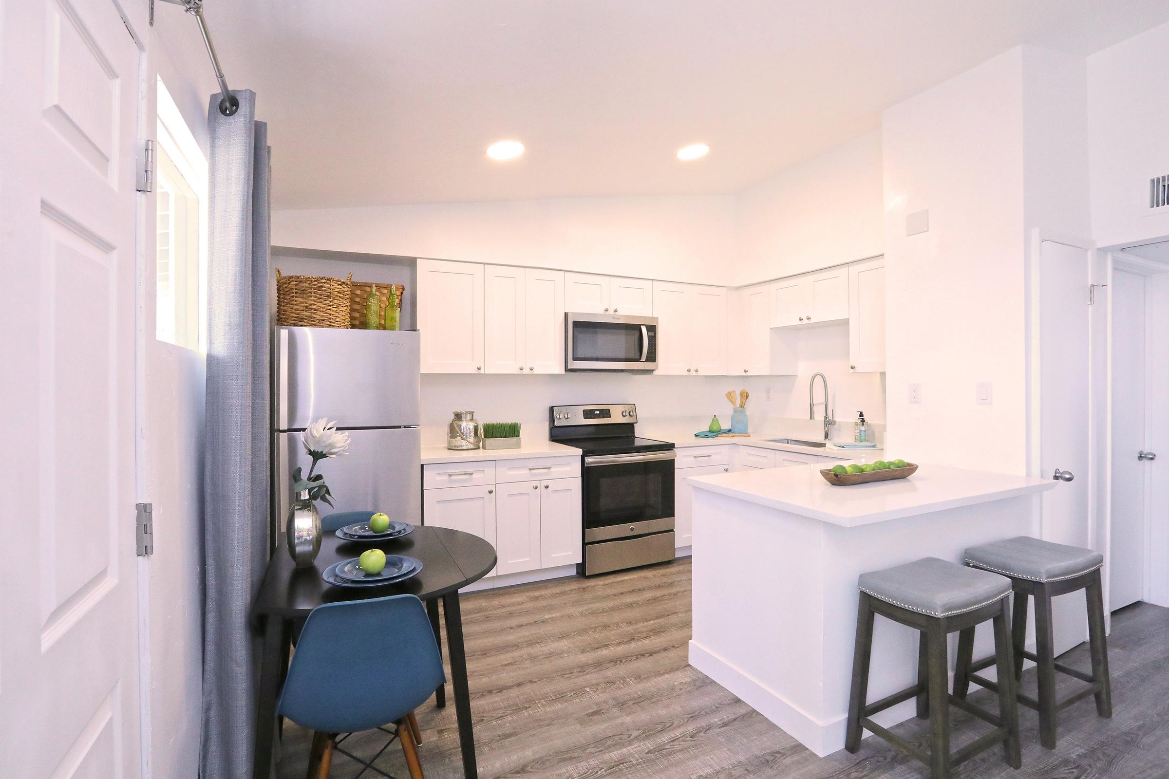 Modern kitchen with white cabinetry and stainless steel appliances. Features a sleek countertop with a sink, a dining table set for two with green plates, and stools at the kitchen island. Bright and airy atmosphere with light-colored walls and gray flooring. Natural light flows into the space through large windows.