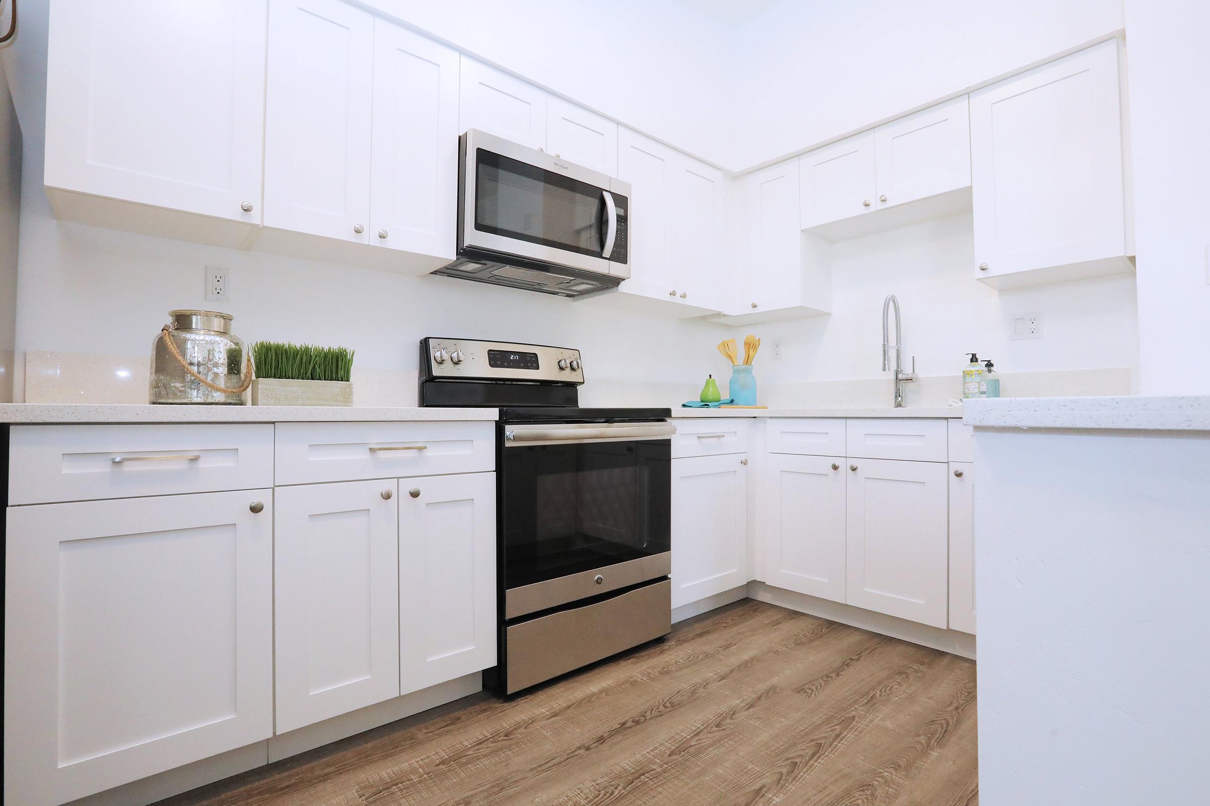 Modern kitchen featuring white cabinetry, stainless steel appliances including a microwave and oven, and a sleek countertop. The floor is a light wood finish, and there's a small decorative arrangement of green grass in a vase. Clean and bright aesthetics, with a focus on functionality and style.