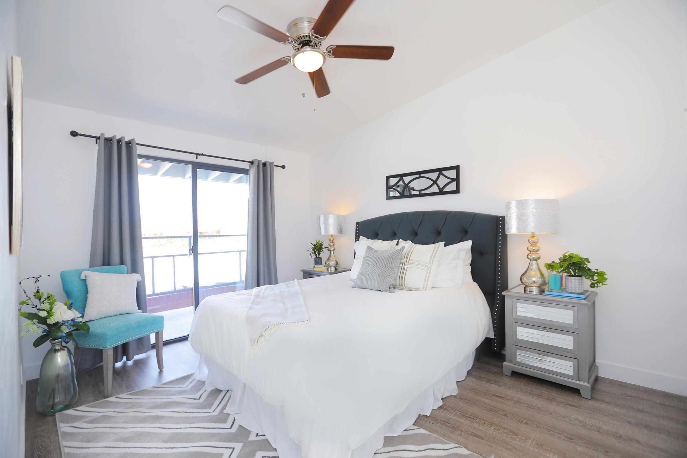 A cozy bedroom featuring a queen-sized bed with white bedding and decorative pillows. There are two bedside tables with lamps, a blue accent chair, and a large window with gray curtains. The room has a ceiling fan and plants for added decor, creating a calm and inviting atmosphere.