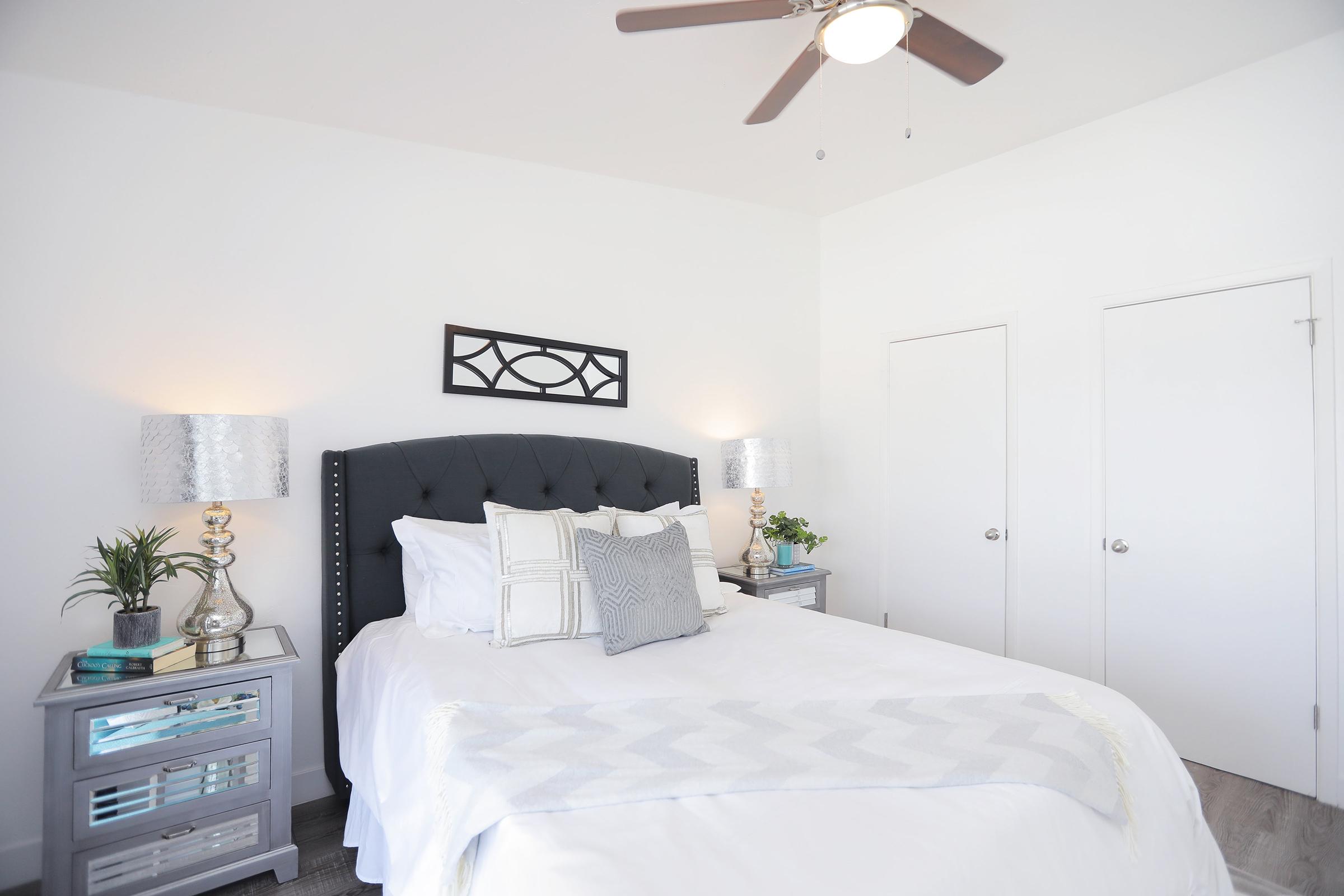 A modern bedroom featuring a queen-sized bed with a dark headboard, white linens, and decorative pillows. There are two bedside tables with lamps and plants, and a geometric wall art piece above the bed. The room has light walls, a ceiling fan, and two closed doors.