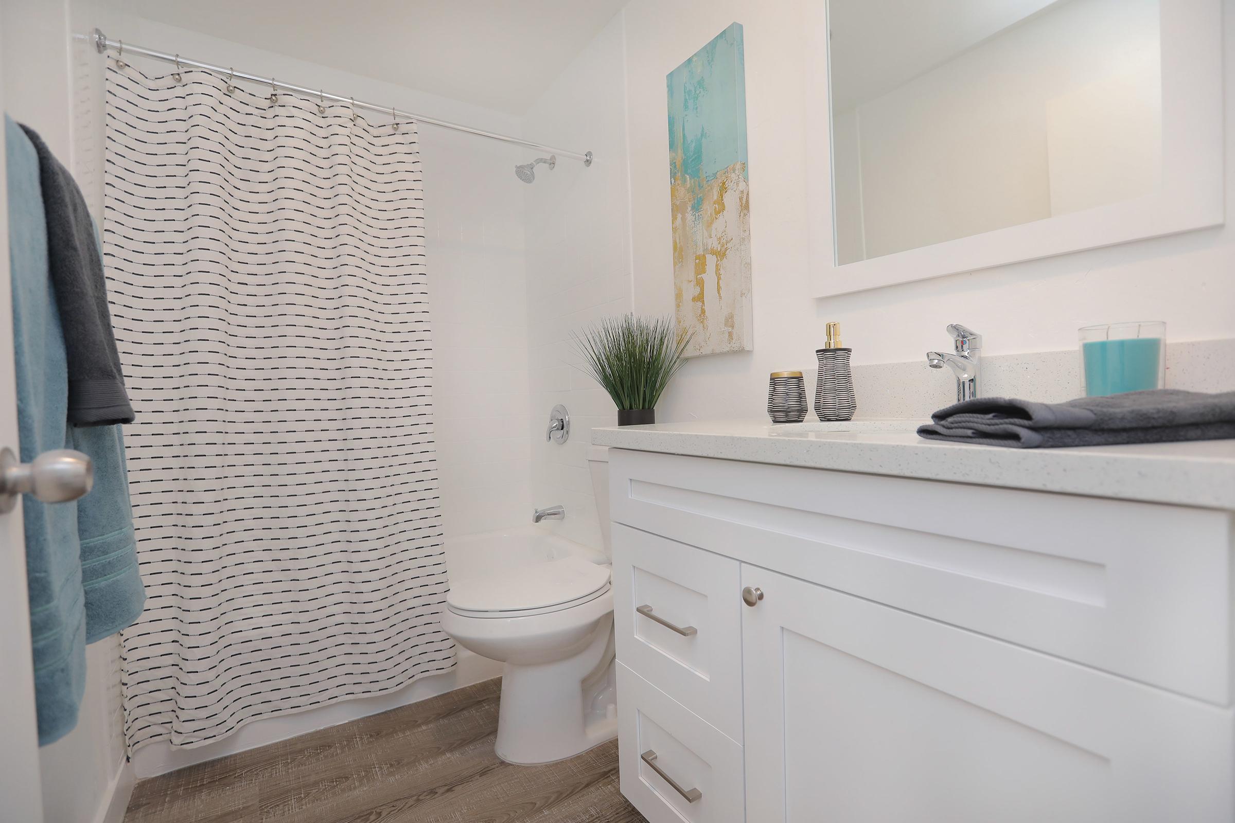 A modern bathroom featuring a white vanity with sink, a shower with a striped curtain, a toilet, and decorative items including a plant, toiletries, and a blue glass. The walls are light-colored, and there’s artwork on the wall, enhancing the clean and contemporary aesthetic of the space.