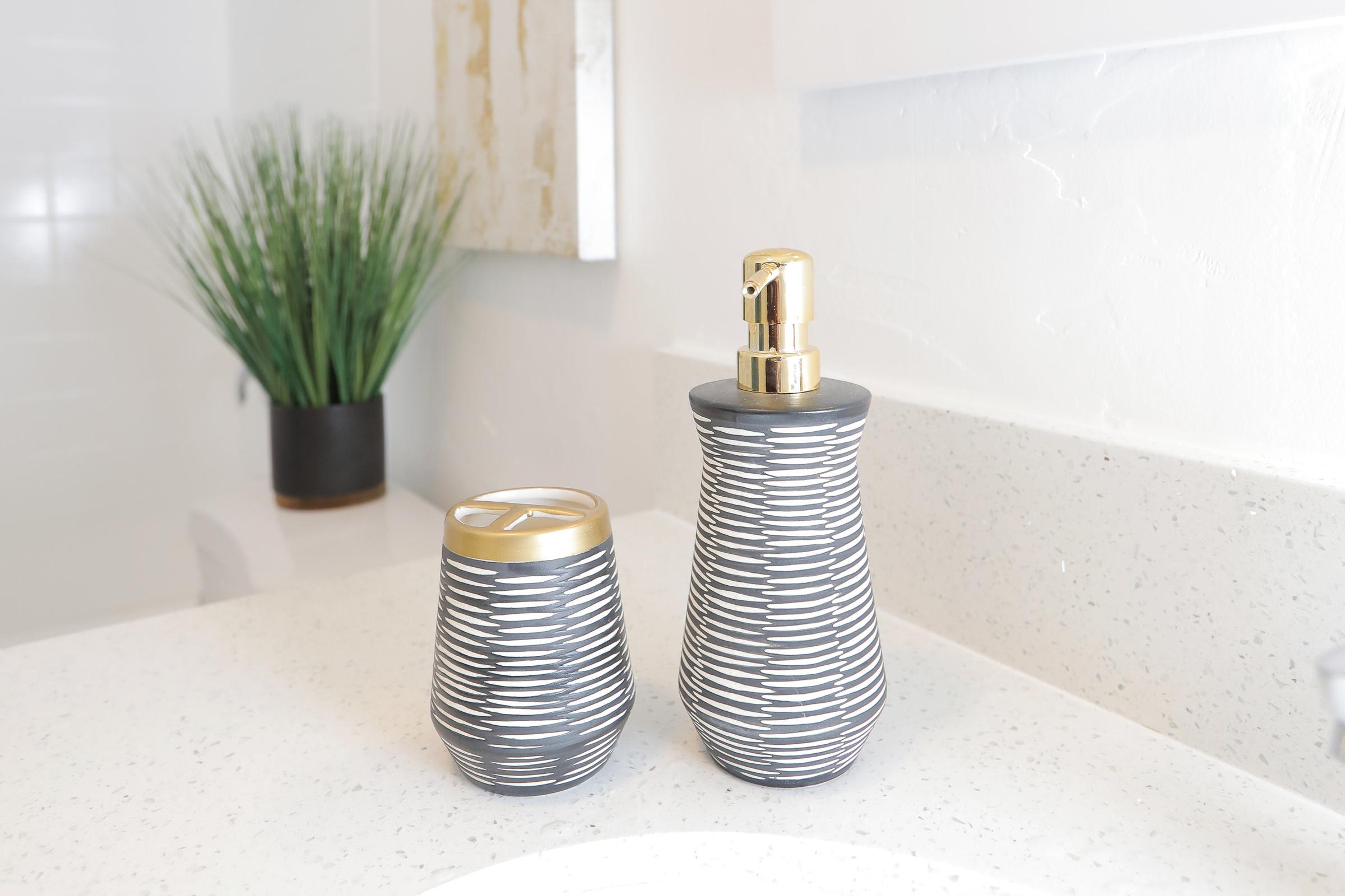 A bathroom countertop featuring a gray and white striped soap dispenser and matching toothbrush holder. In the background, there is a small potted plant adding a touch of greenery to the serene setting. The surface is a light-colored speckled countertop, enhancing the modern aesthetic.