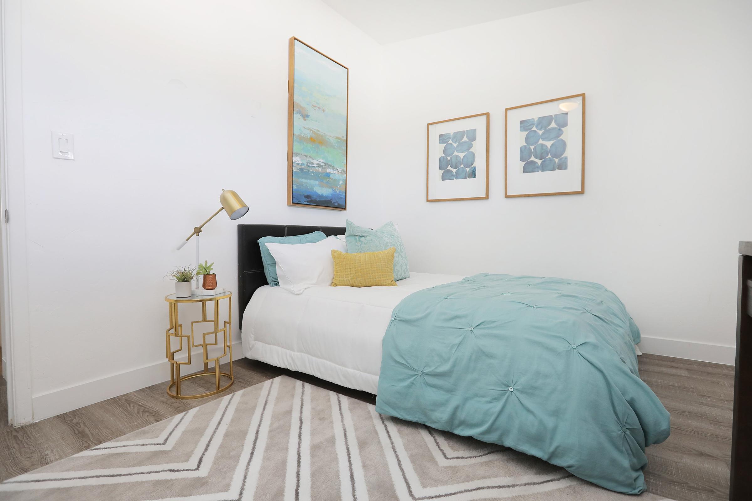 A cozy bedroom featuring a single bed with a light blue comforter, decorative pillows, and a yellow accent pillow. A small nightstand with a plant and a lamp is beside the bed. The walls are adorned with two framed artworks, and a stylish rug covers the floor, contributing to a modern and inviting atmosphere.