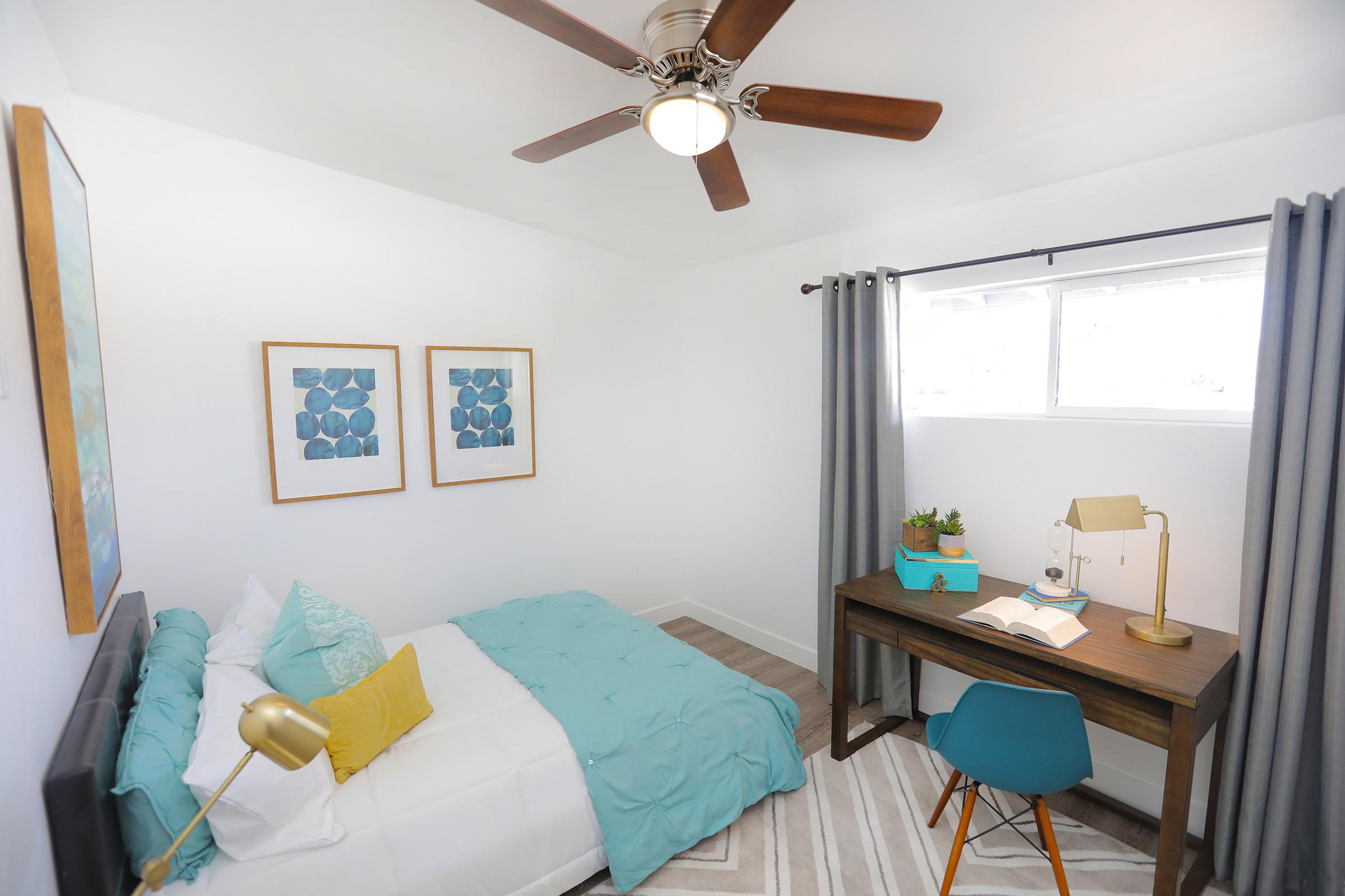A cozy bedroom featuring a light blue over-blanket on a bed with white linens, two framed artworks on the walls, a wooden desk with a lamp and a book, a modern blue chair, and a ceiling fan. Natural light streams in through a window with gray curtains, creating a bright and inviting atmosphere.